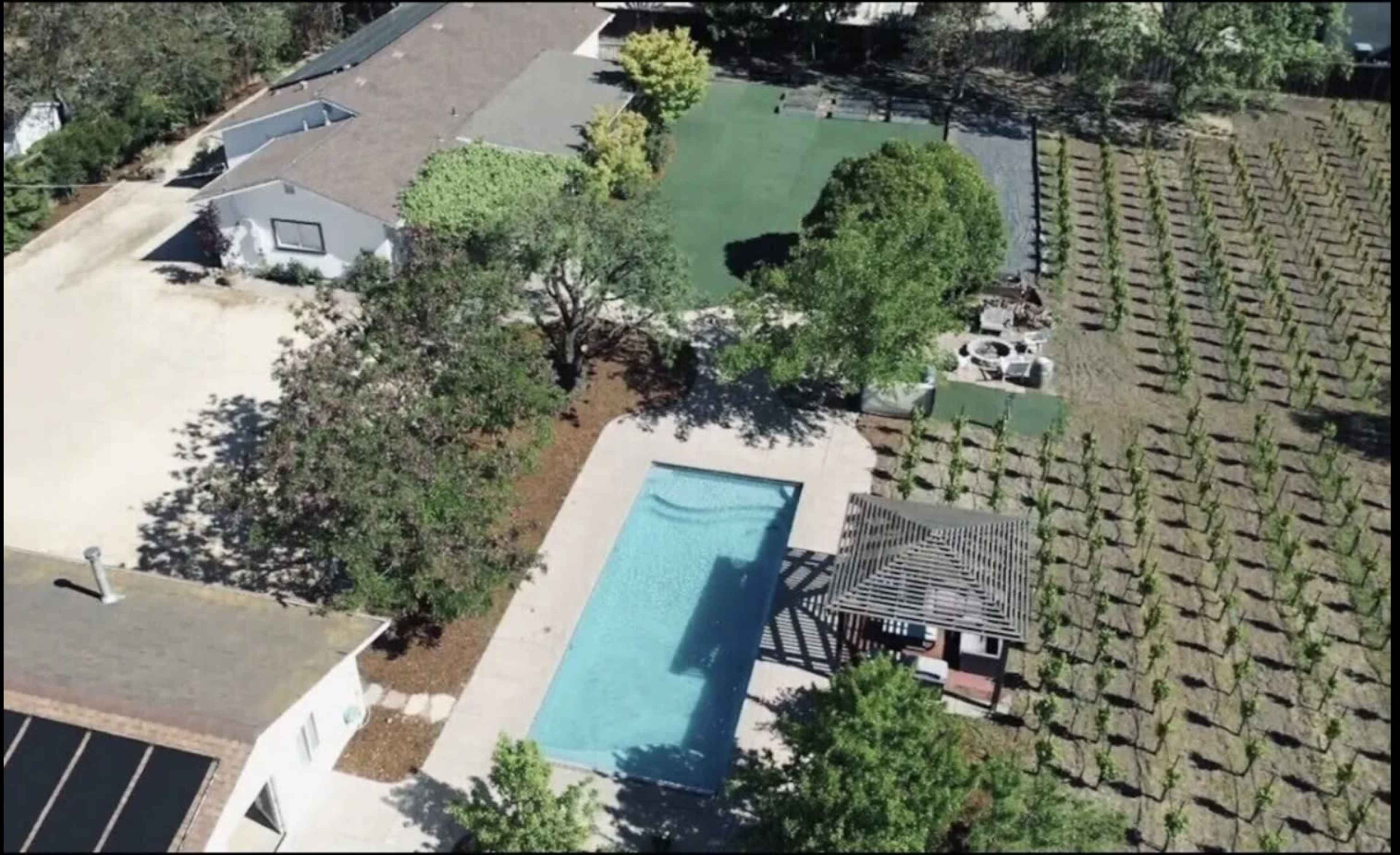 An aerial view of a property featuring a swimming pool, a landscaped yard with trees, and a vineyard.