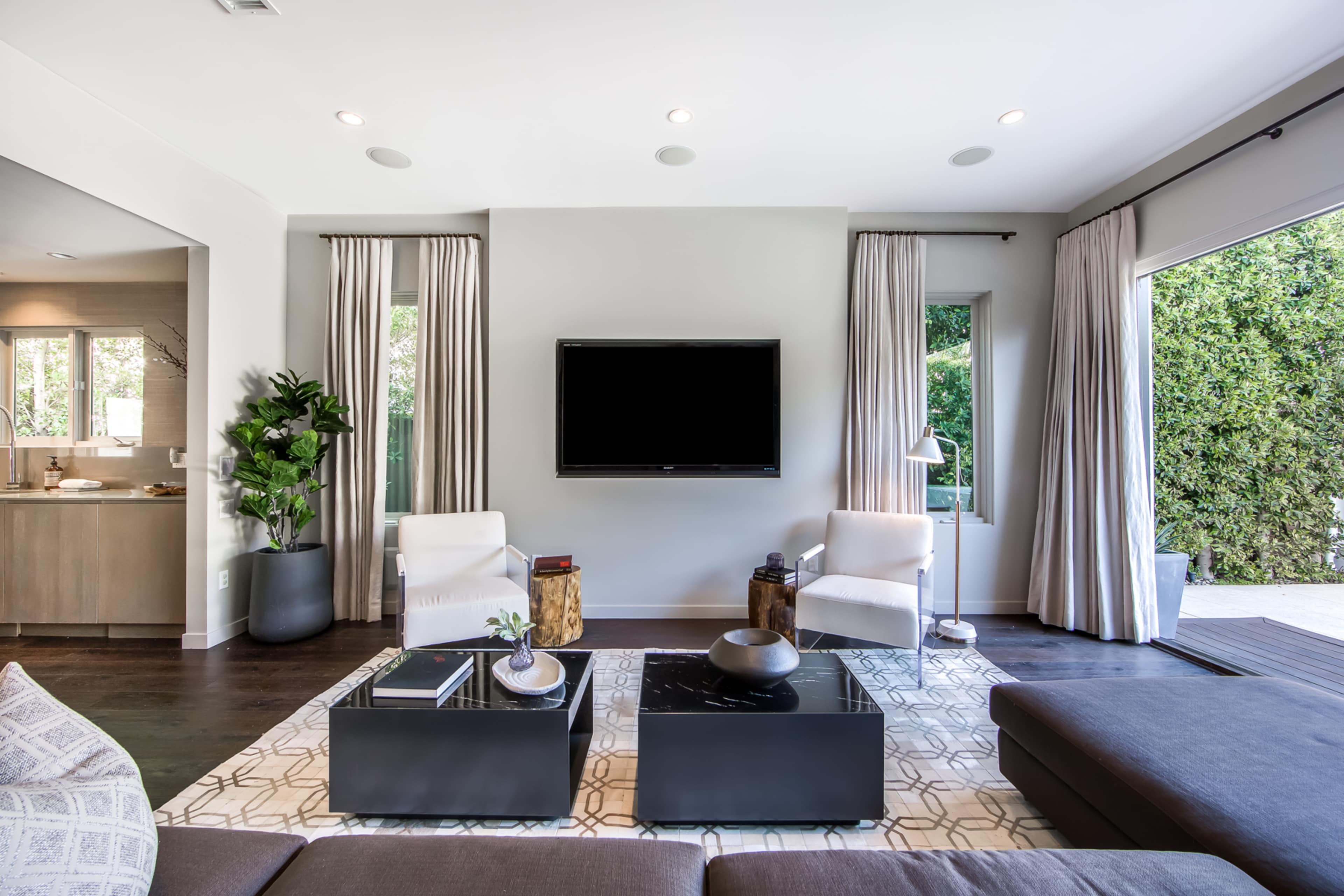 The living room features a television mounted on the wall, two white armchairs, a dark coffee table, and large windows that open to a patio.