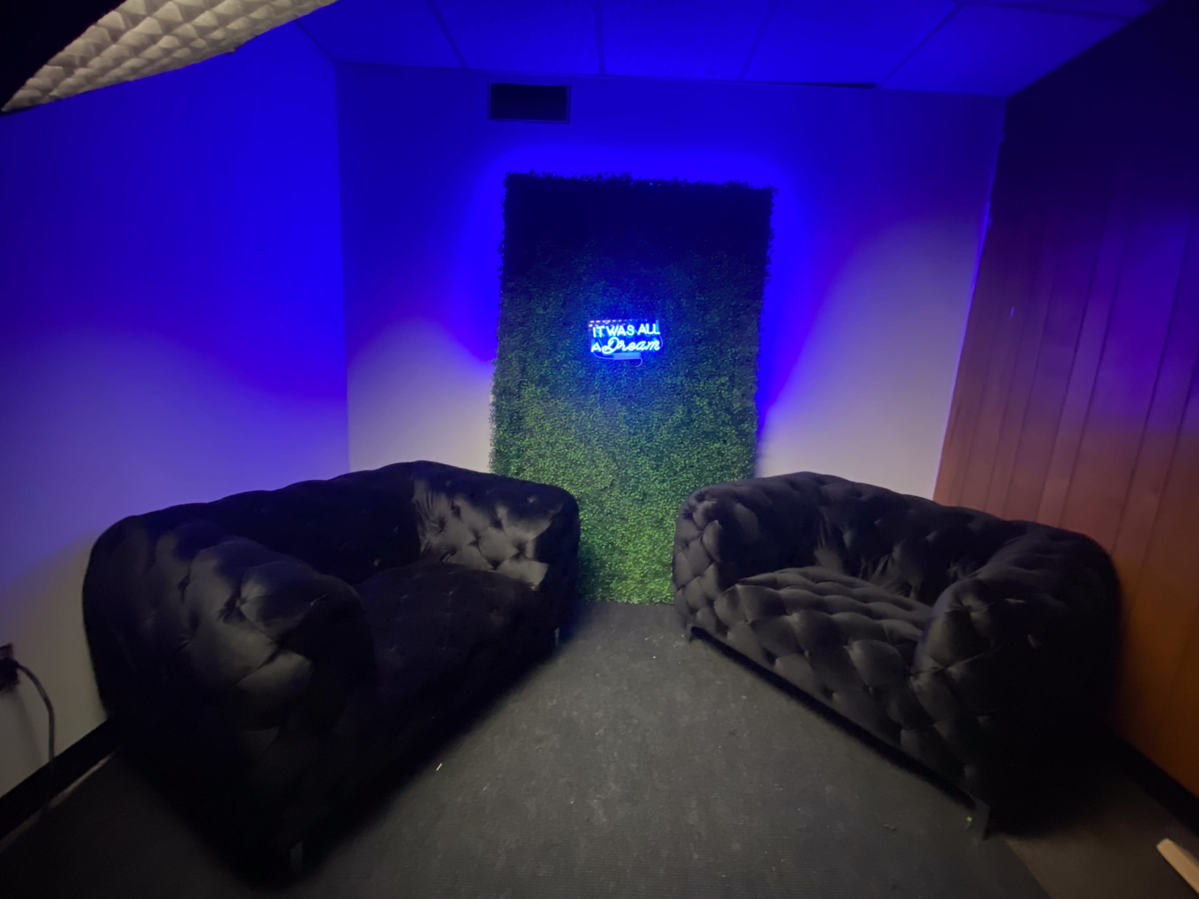 The image shows a small lounge area with two black tufted sofas facing a green wall with a neon sign that reads "It was all a dream."