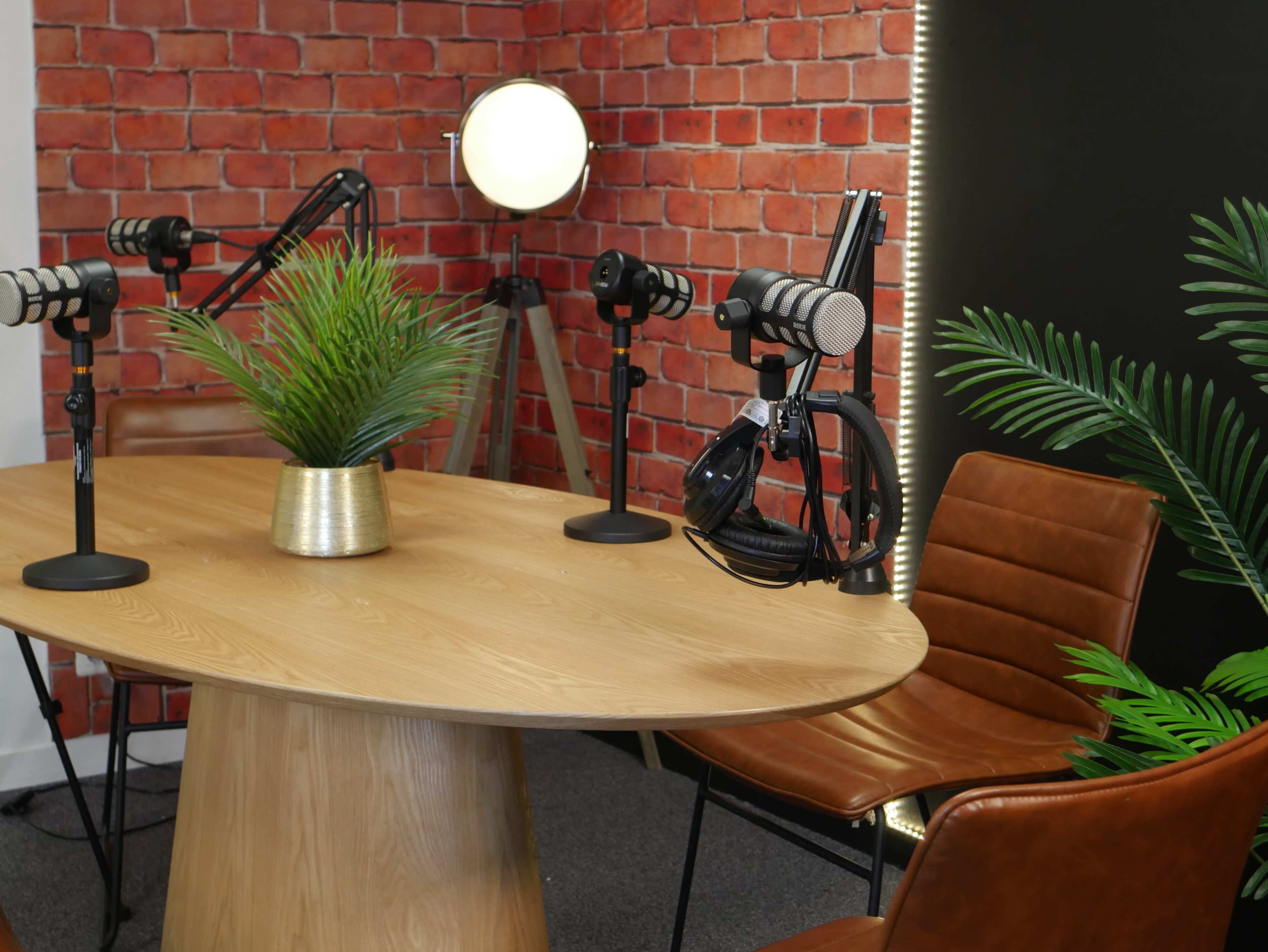 The image shows a podcast recording setup featuring a round wooden table, three microphone stands, a potted plant, and two brown leather chairs against a brick wall background.