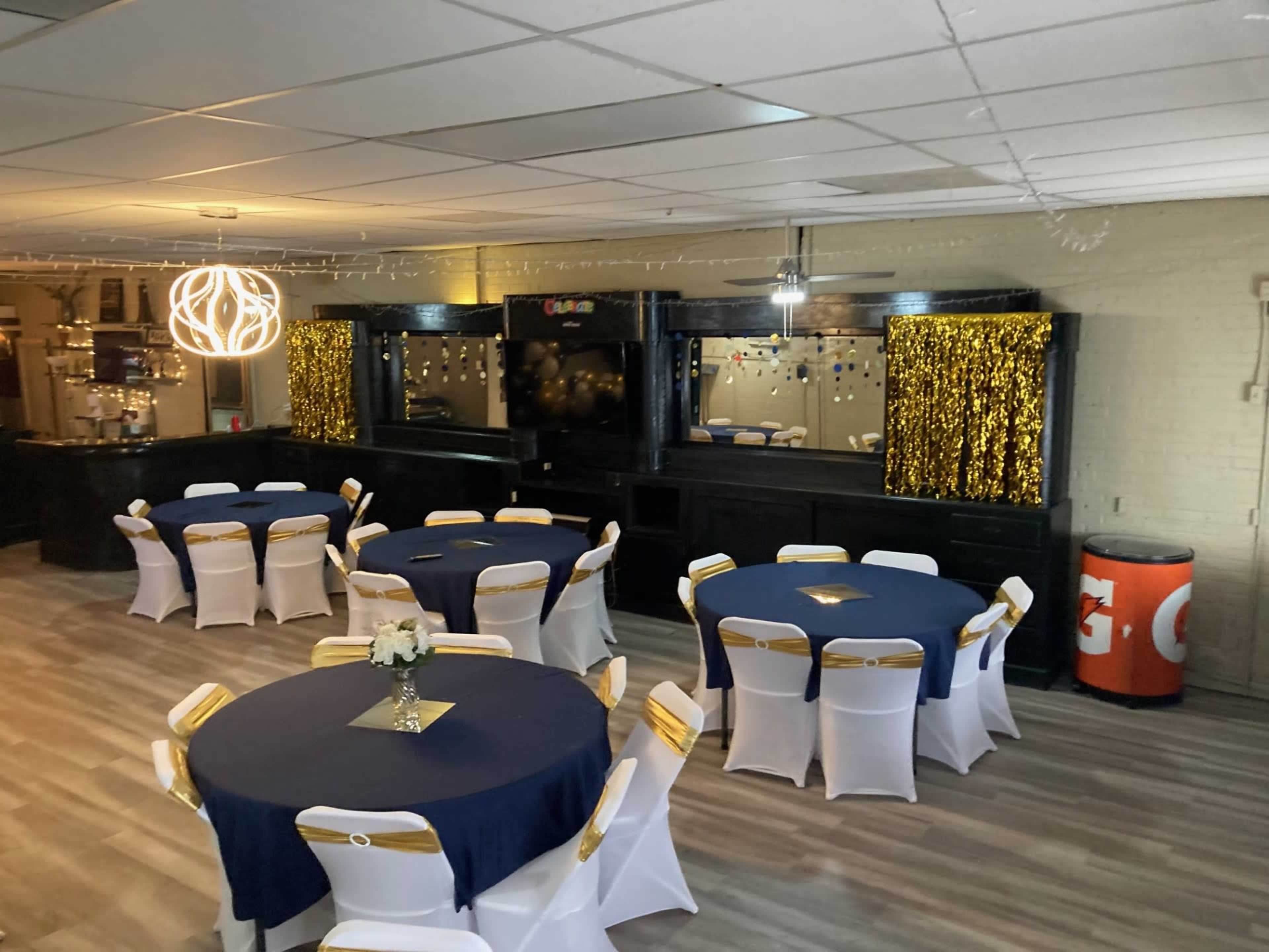 A decorated event space with four round tables covered in blue tablecloths and white chairs, featuring gold ribbon accents, and a backdrop with decorative elements on the wall.