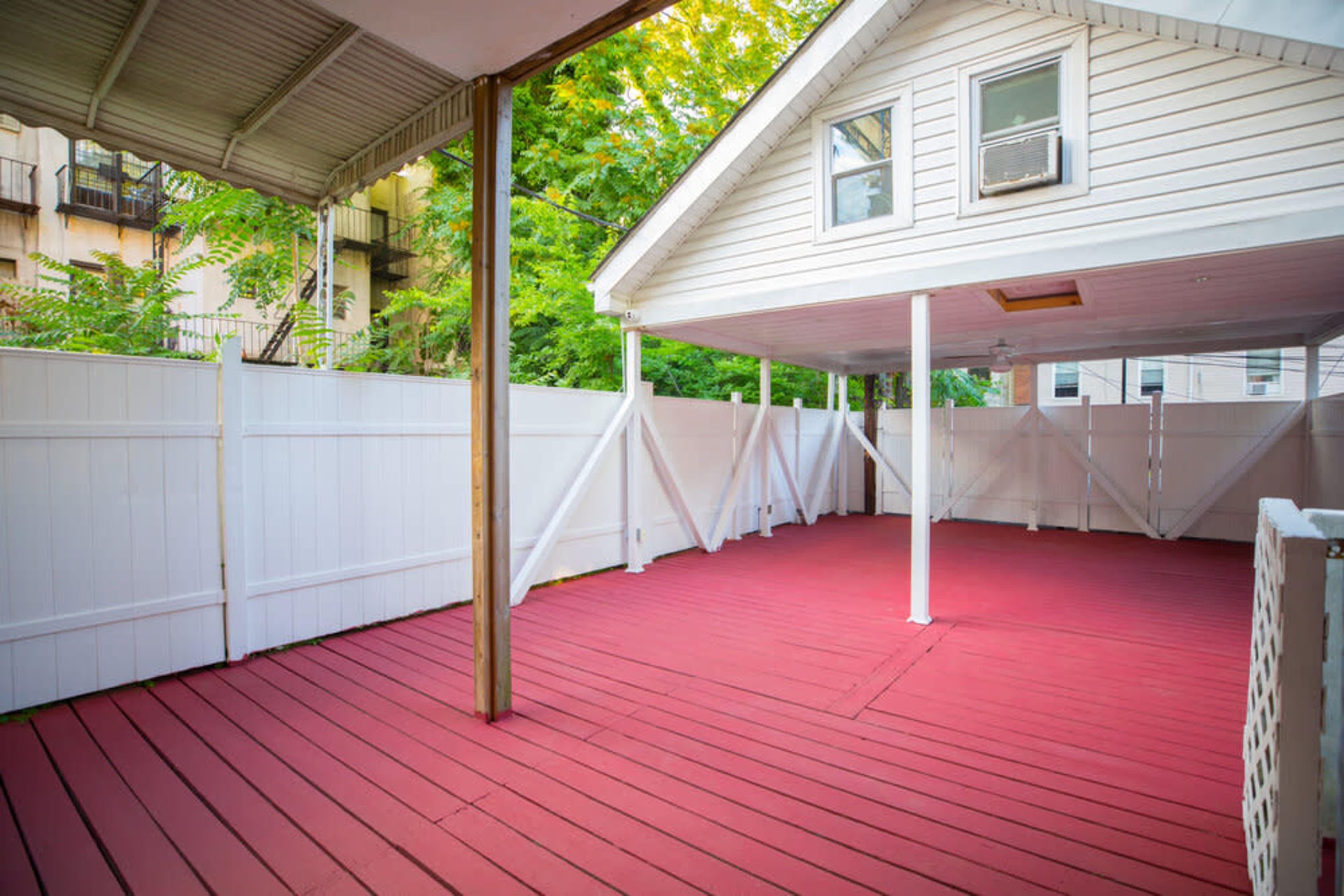 A covered outdoor space with a red wooden floor and white privacy fencing, surrounded by greenery.