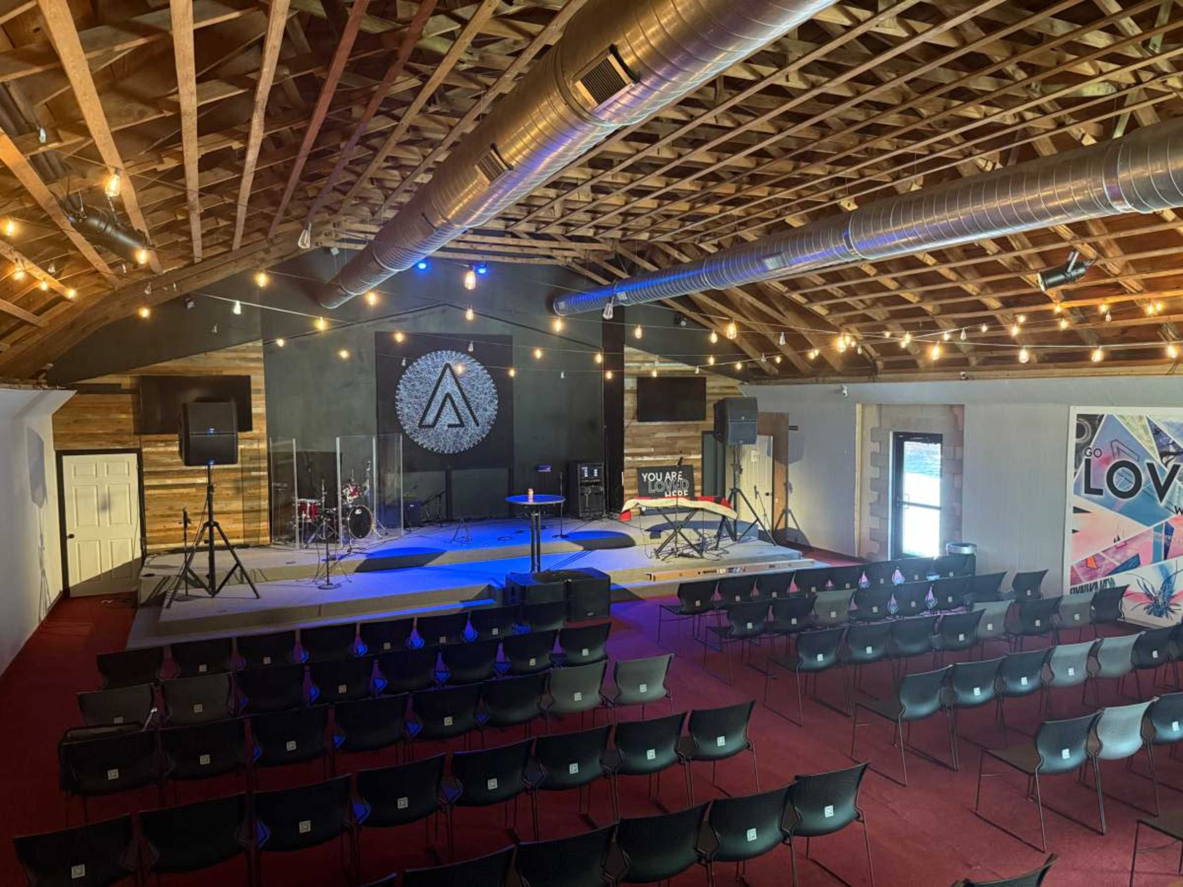 The image shows an indoor space set up for an event, featuring rows of black chairs facing a stage with musical equipment, under a ceiling with exposed beams and string lights.