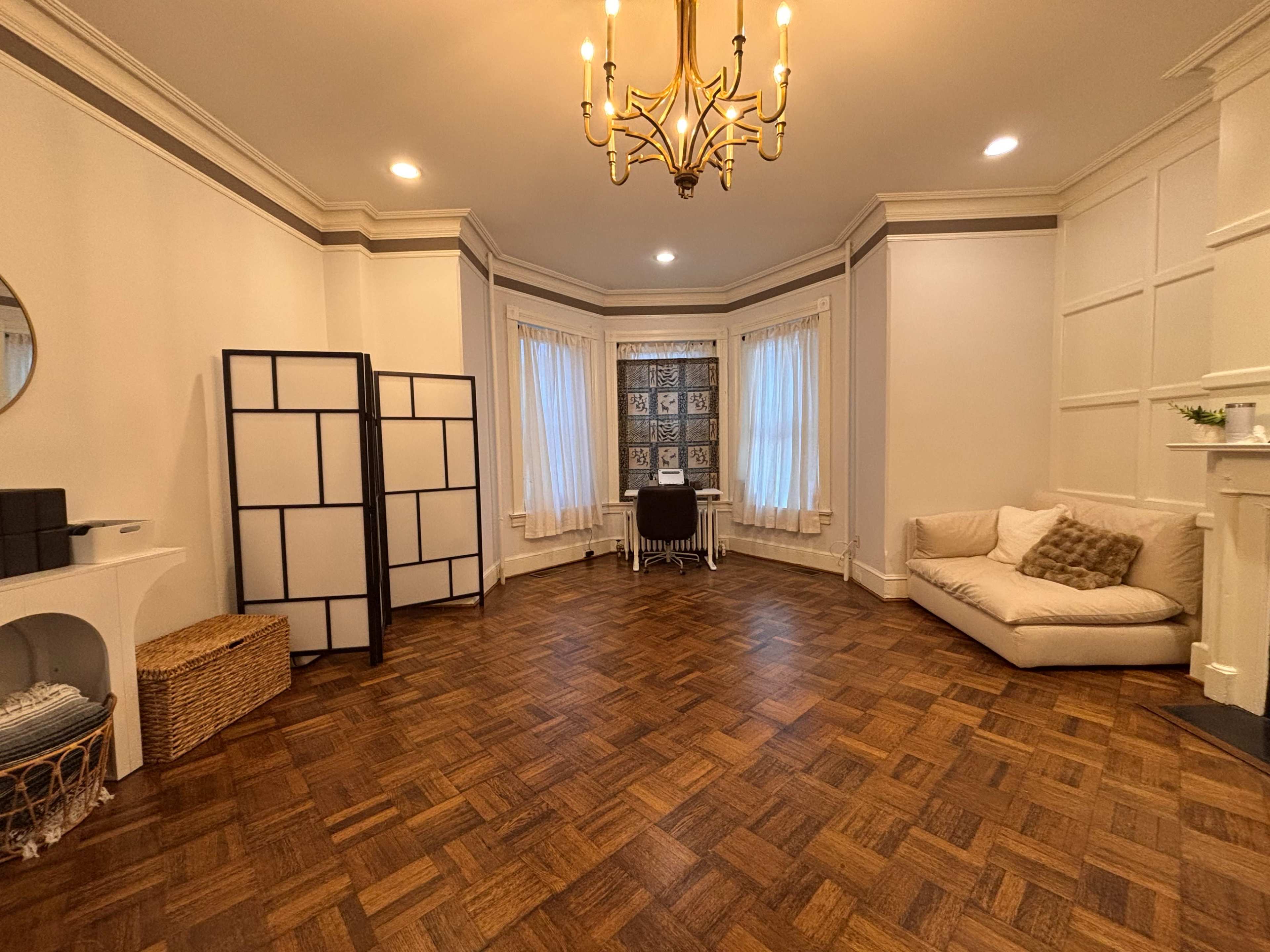 The image shows a spacious room with wooden floors, bright windows, a large chandelier, a sectional sofa, a folding screen, and a table set against a patterned backdrop.