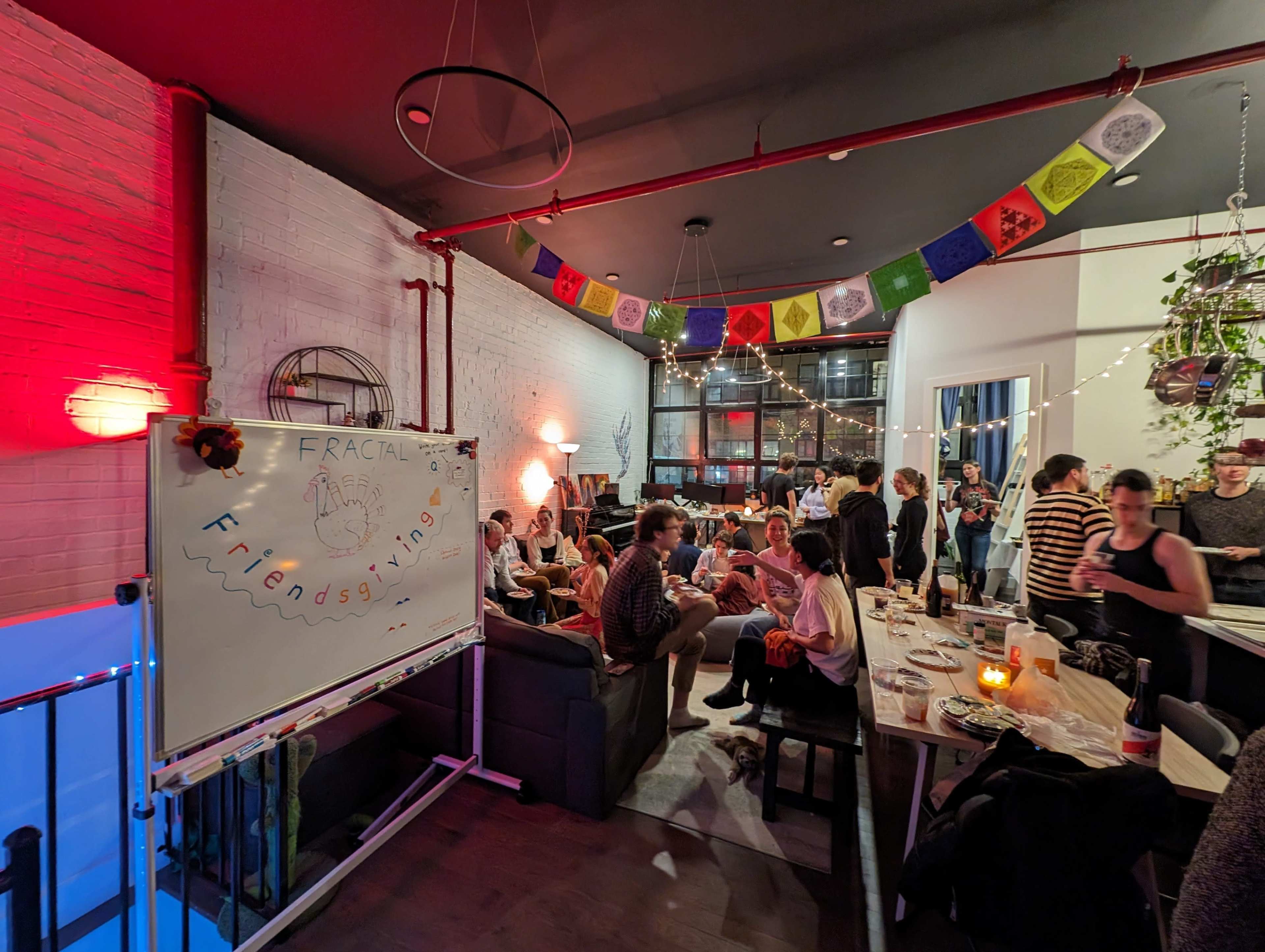 A lively gathering is taking place in a room decorated with colorful prayer flags, where people are seated at tables and conversing.