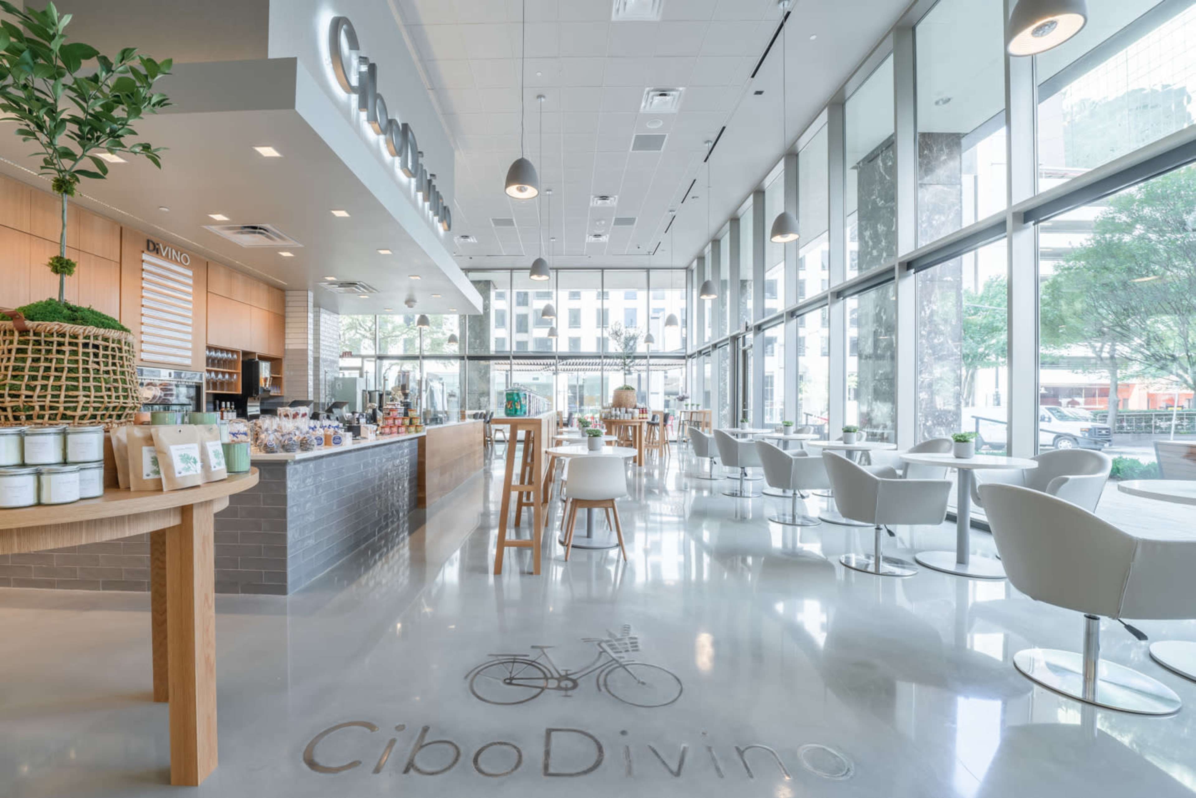 The image shows a modern café interior with large windows, a wooden counter displaying various products, and white seating arrangements.