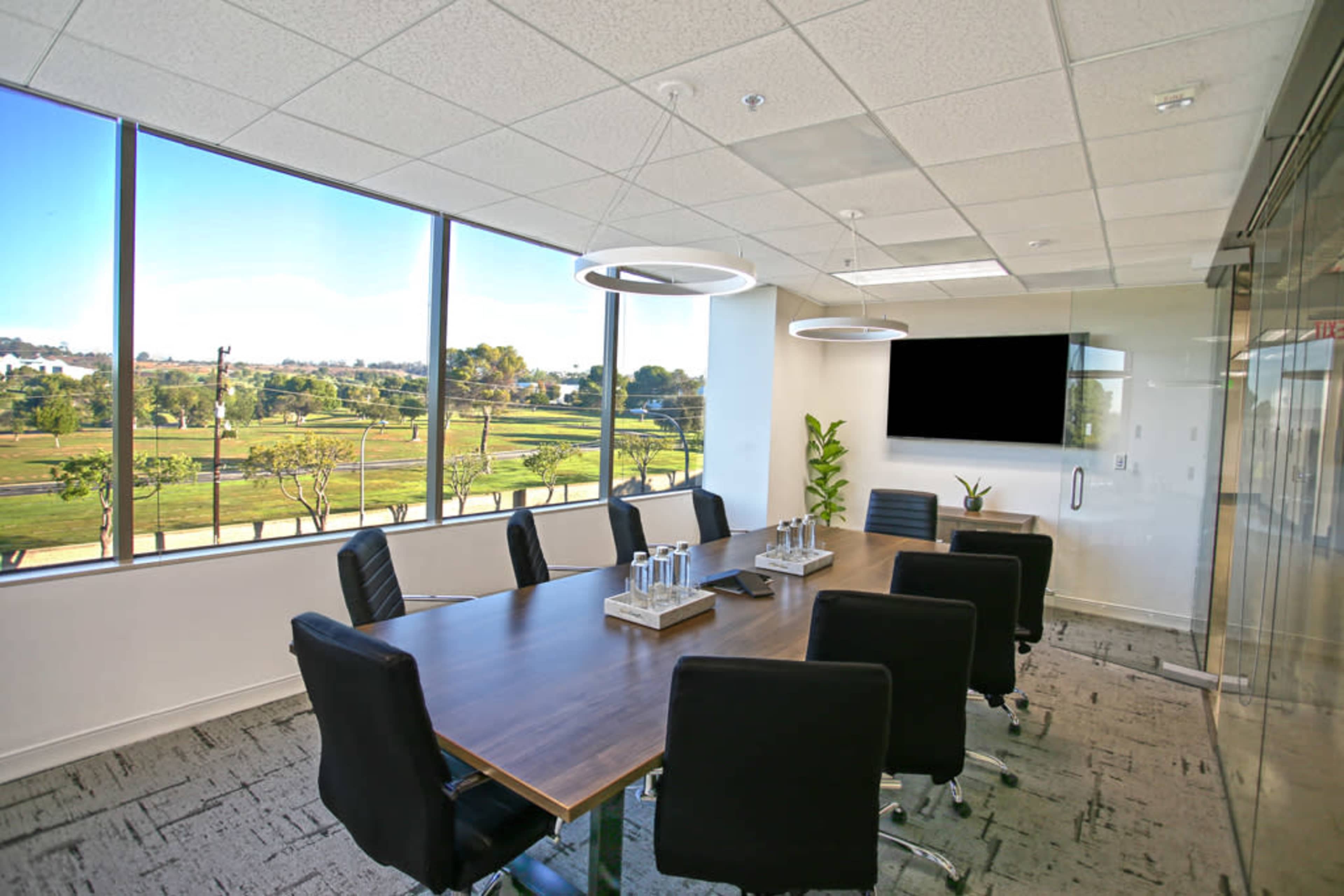 A modern conference room features a large wooden table surrounded by black chairs, with large windows offering a view of greenery outside.