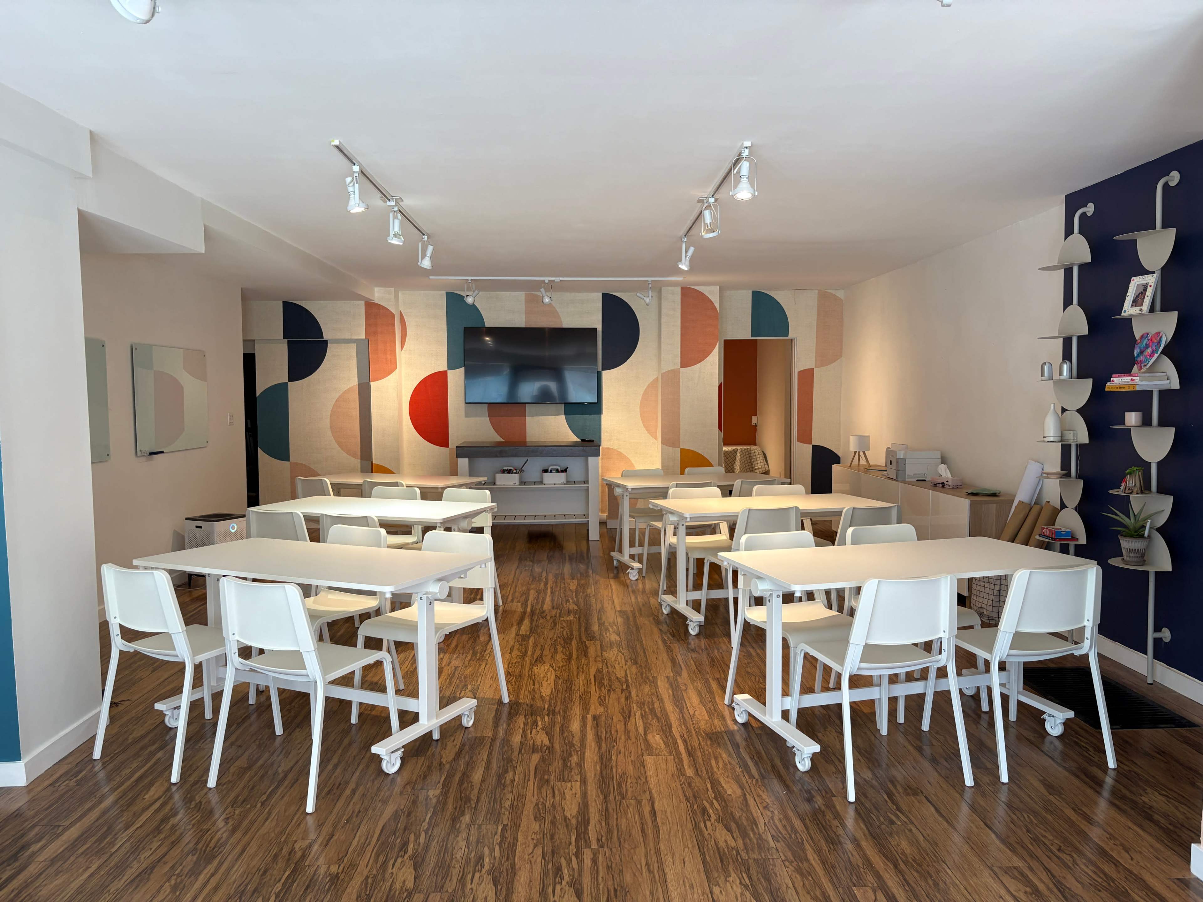 The image shows a modern classroom with multiple white tables arranged in rows, chairs, a wall-mounted television, and a colorful patterned wall.