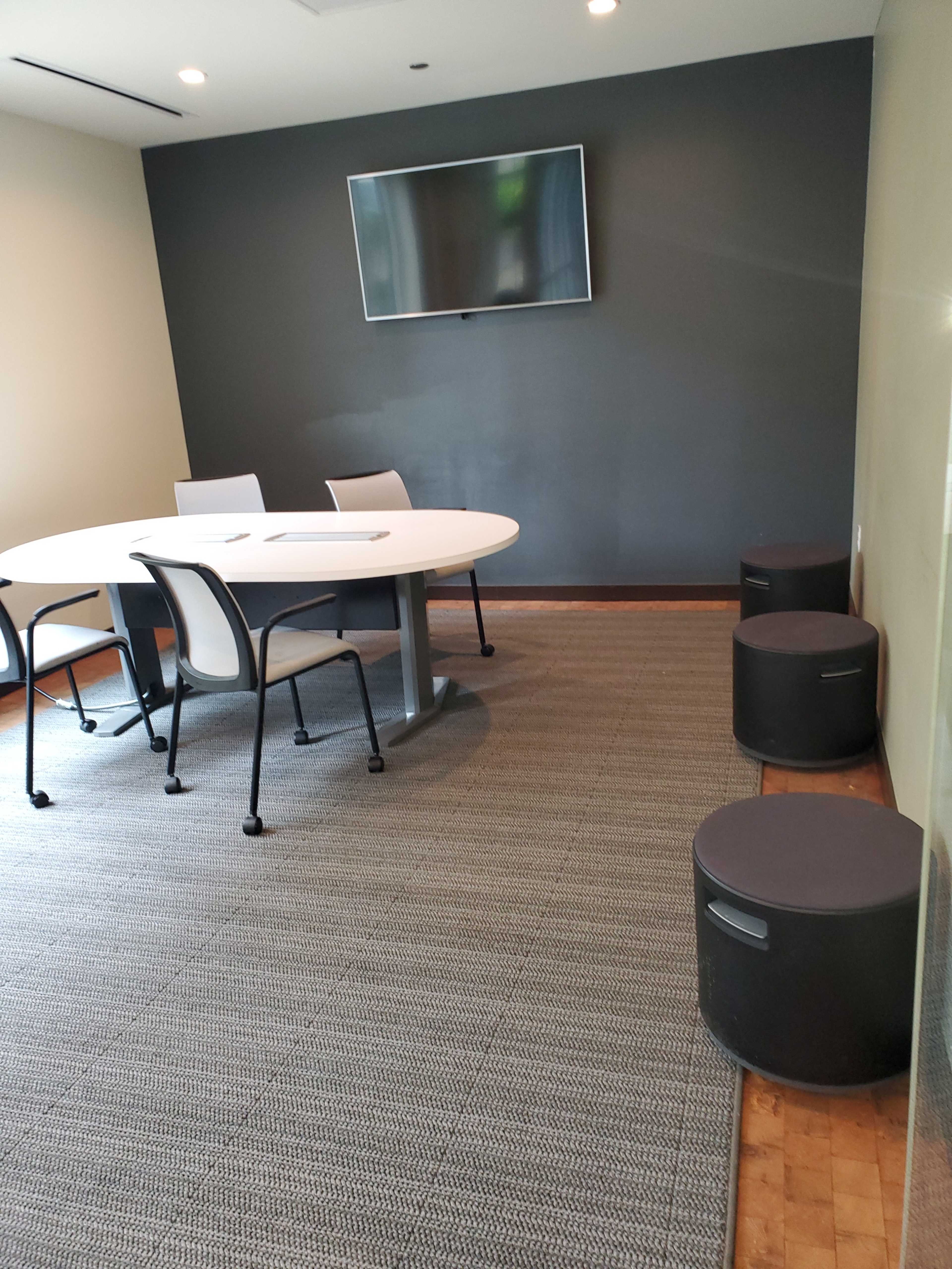 A conference room features a round table surrounded by chairs, with a television mounted on the wall and cylindrical stools positioned nearby.