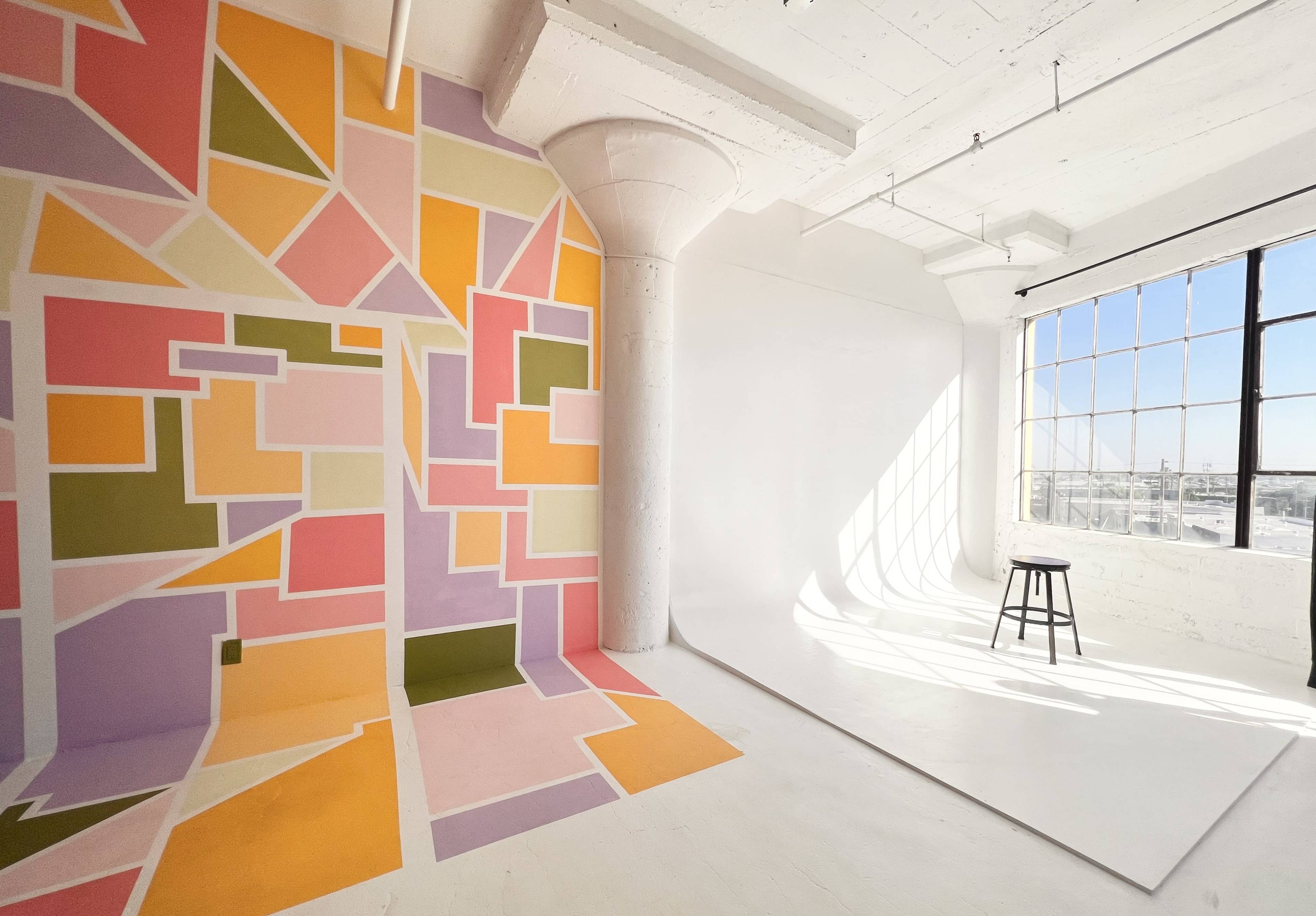 The scene features a bright open studio space with a colorful geometric mural on one wall and a plain white backdrop illuminated by natural light from a large window.