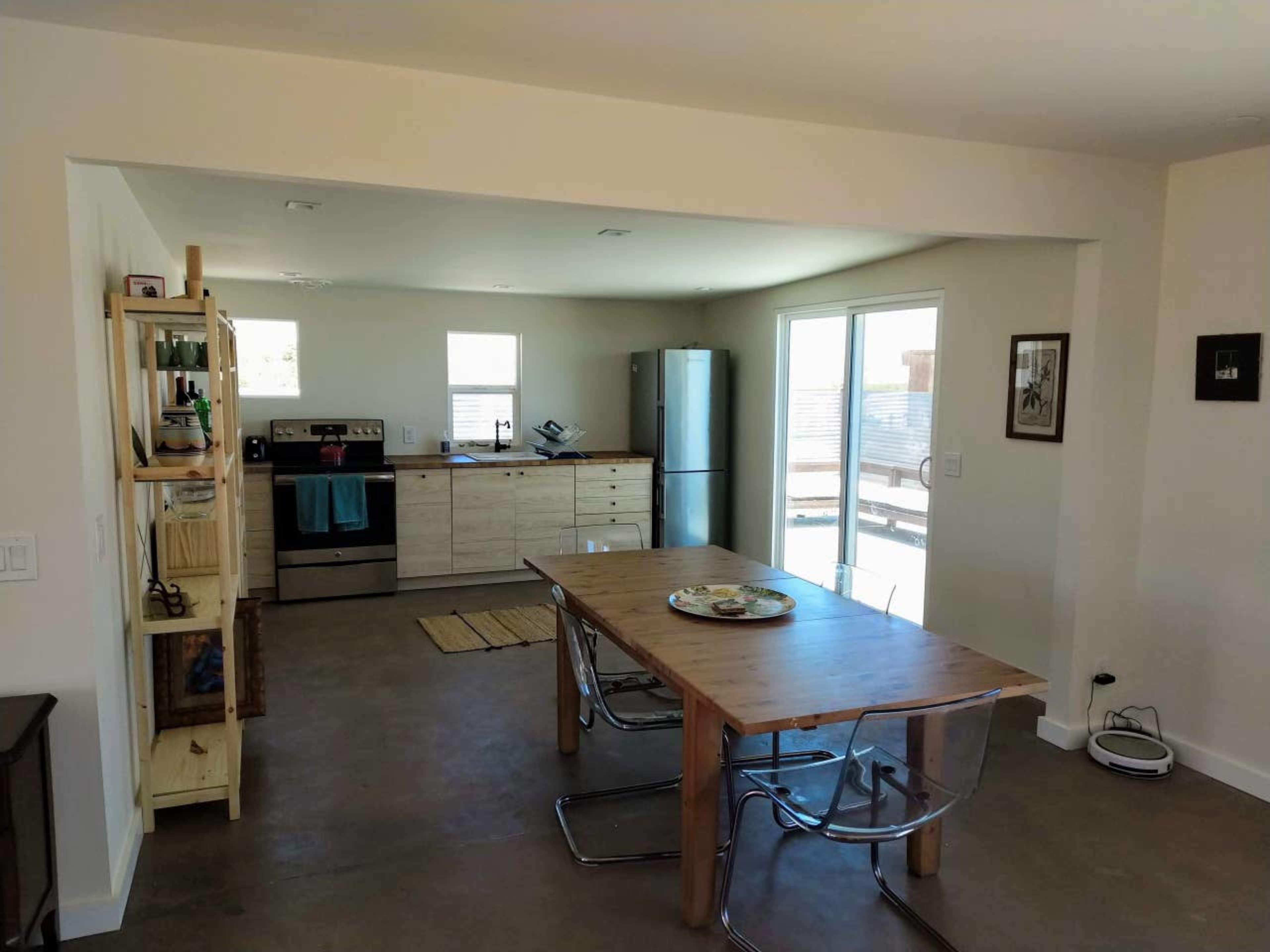 A minimalist kitchen and dining area with a wooden table, stainless steel appliances, and a sliding glass door leading outside.