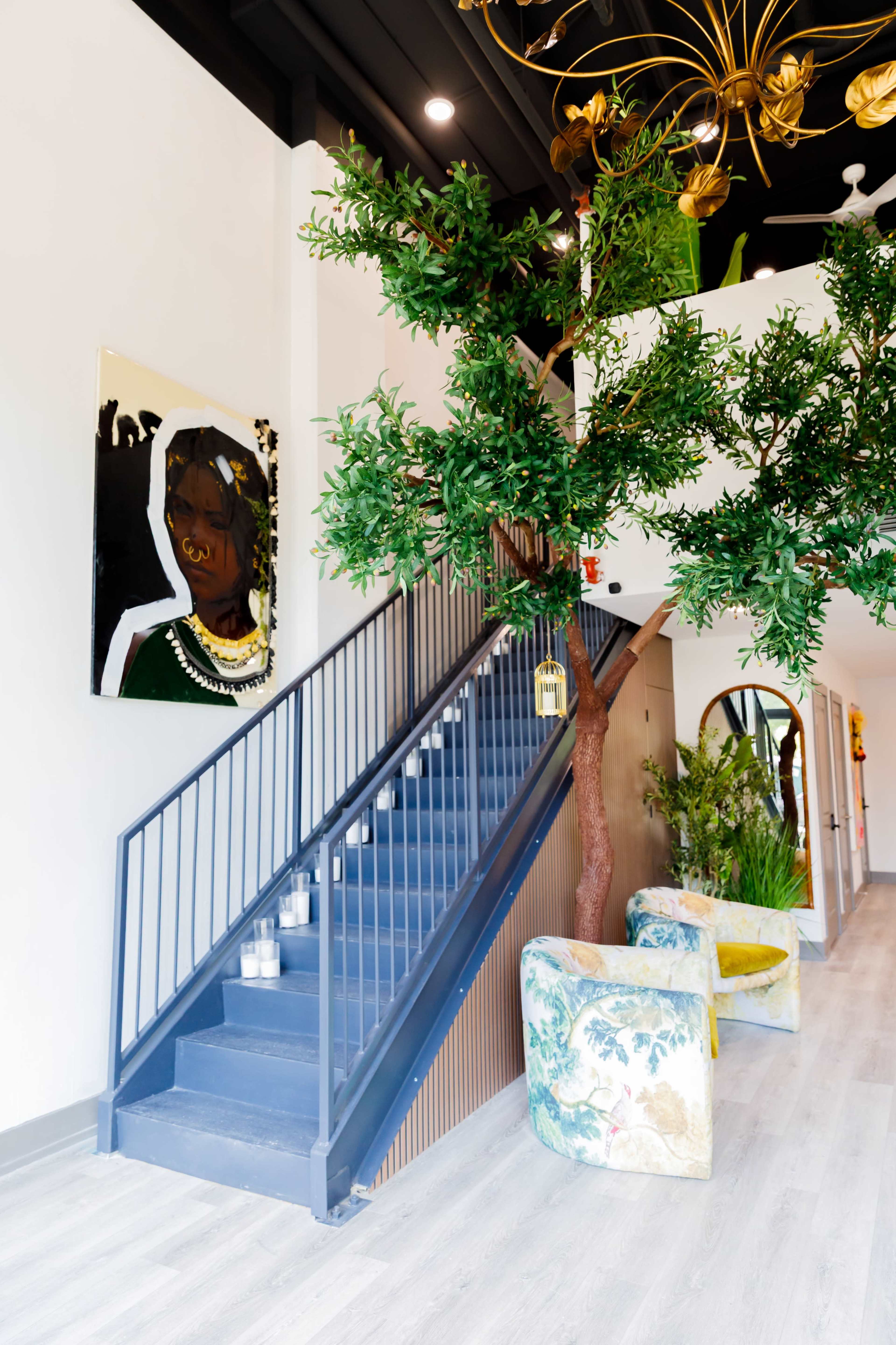 The image shows a modern interior featuring a staircase with a blue railing, flanked by a decorative tree and colorful armchairs.