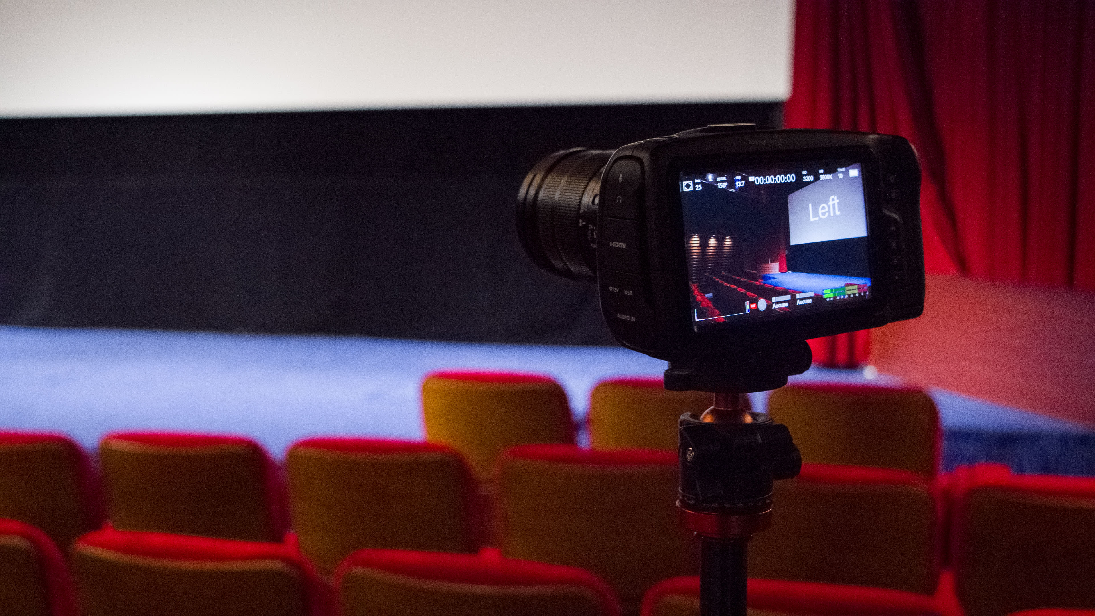 A camera is positioned on a tripod, focused on an empty cinema screen displaying the word "Left," with red theater seats visible in the foreground.