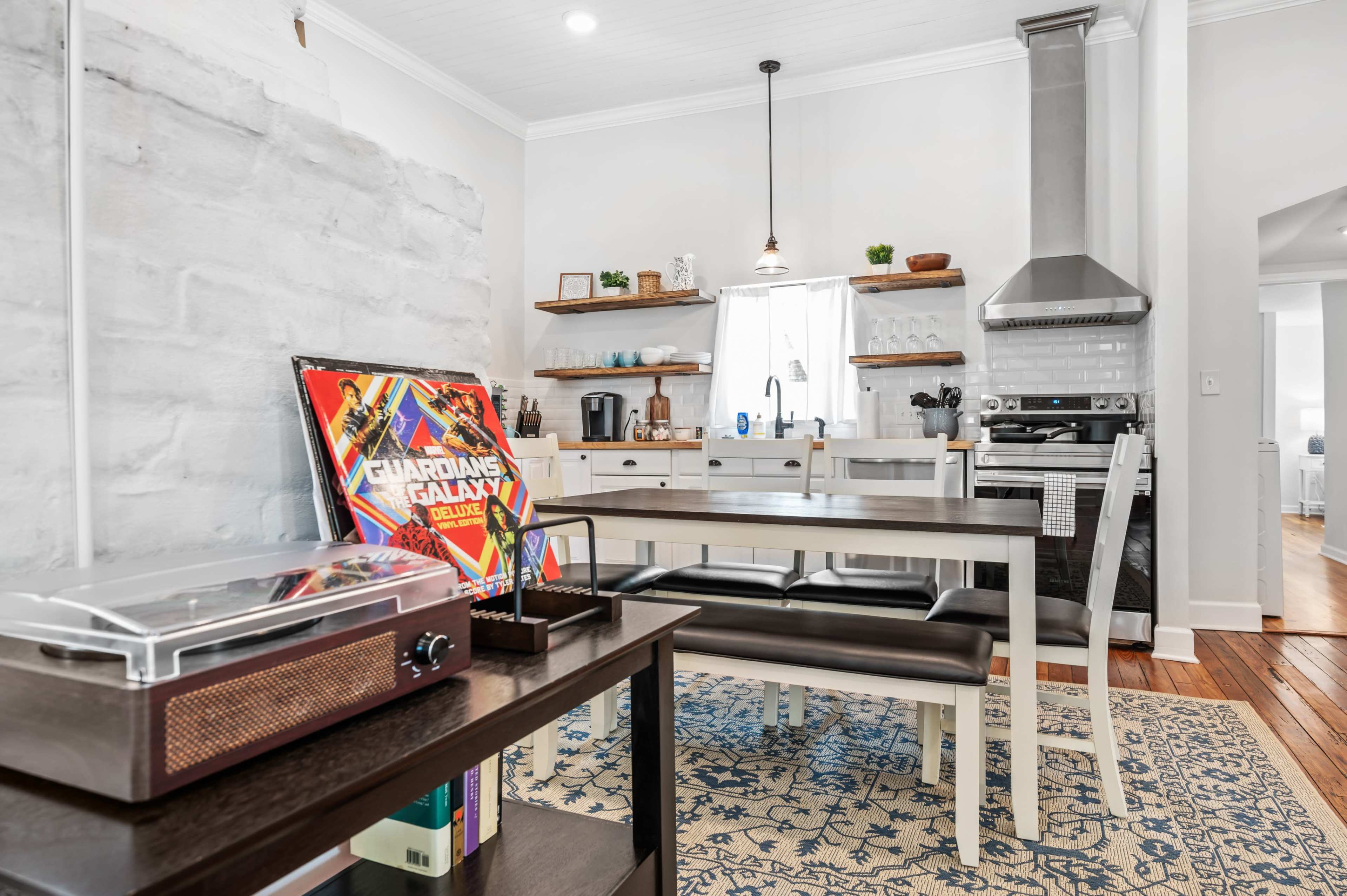 A clean, modern kitchen and dining area features a wooden table with black benches, a record player on a shelf, and stainless steel appliances.