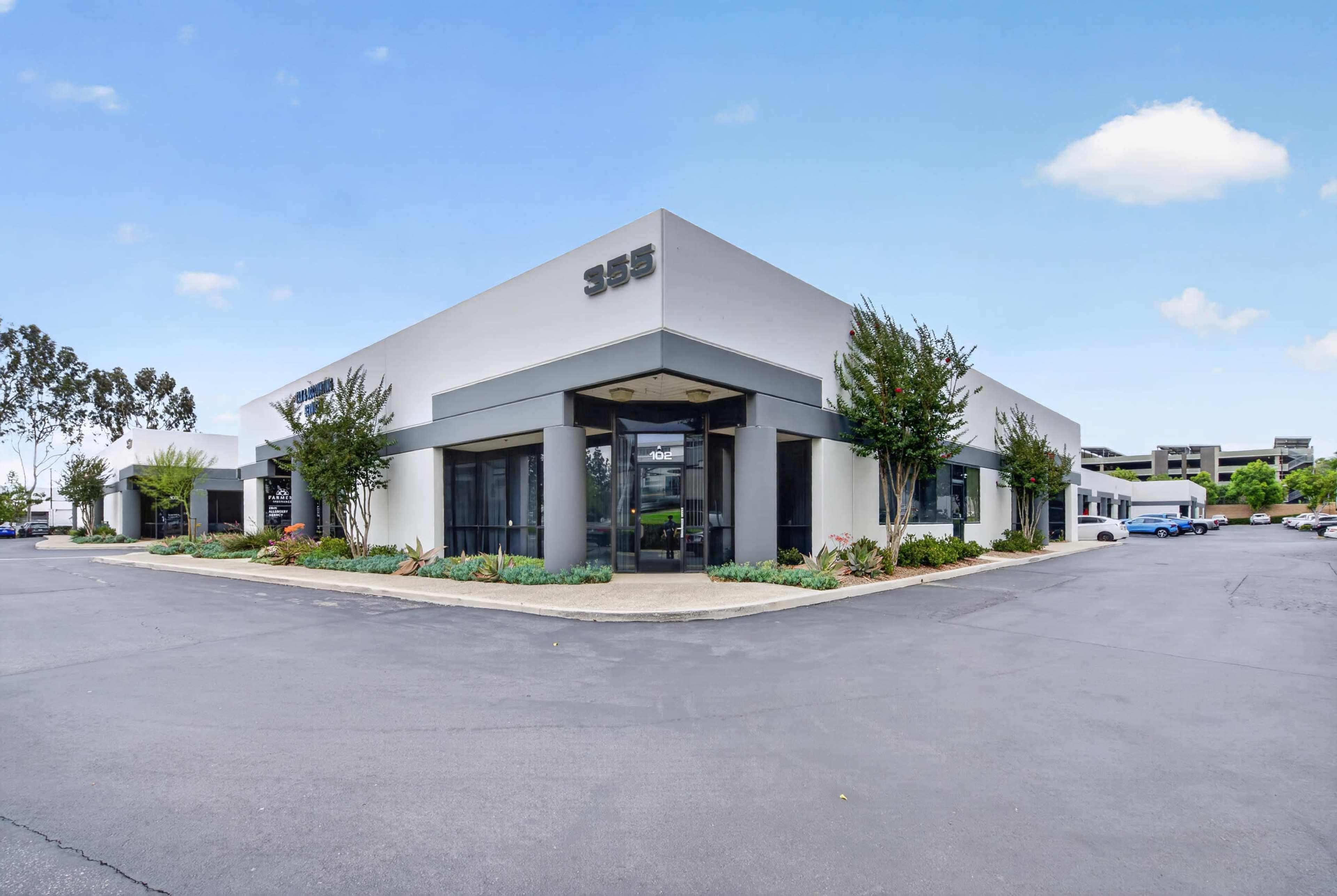 The image shows a modern commercial building with large glass windows and a landscaped parking area.