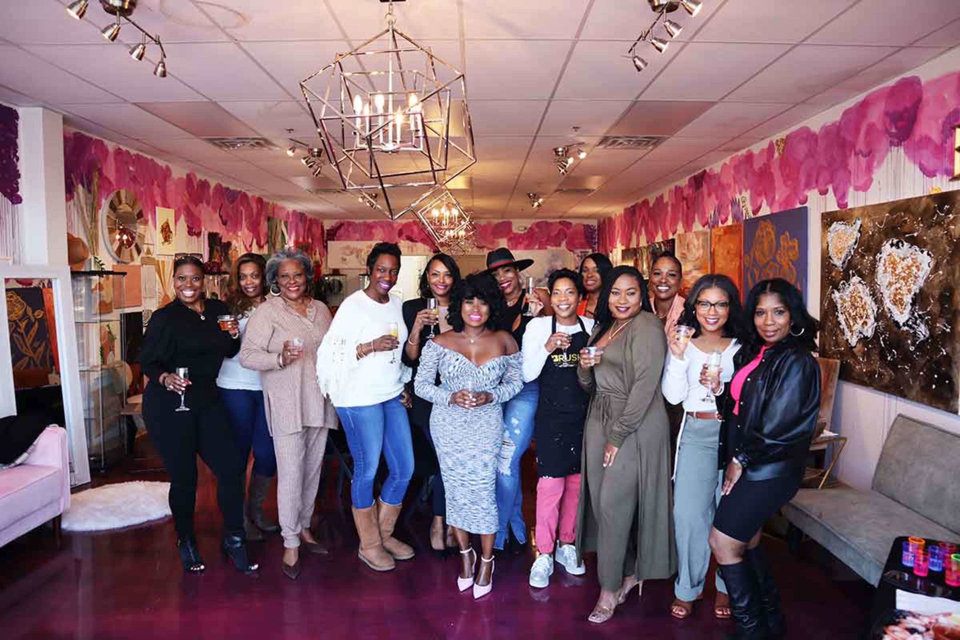 A group of women holds drinks and smiles together in a decorated indoor space with pink accents and artwork.