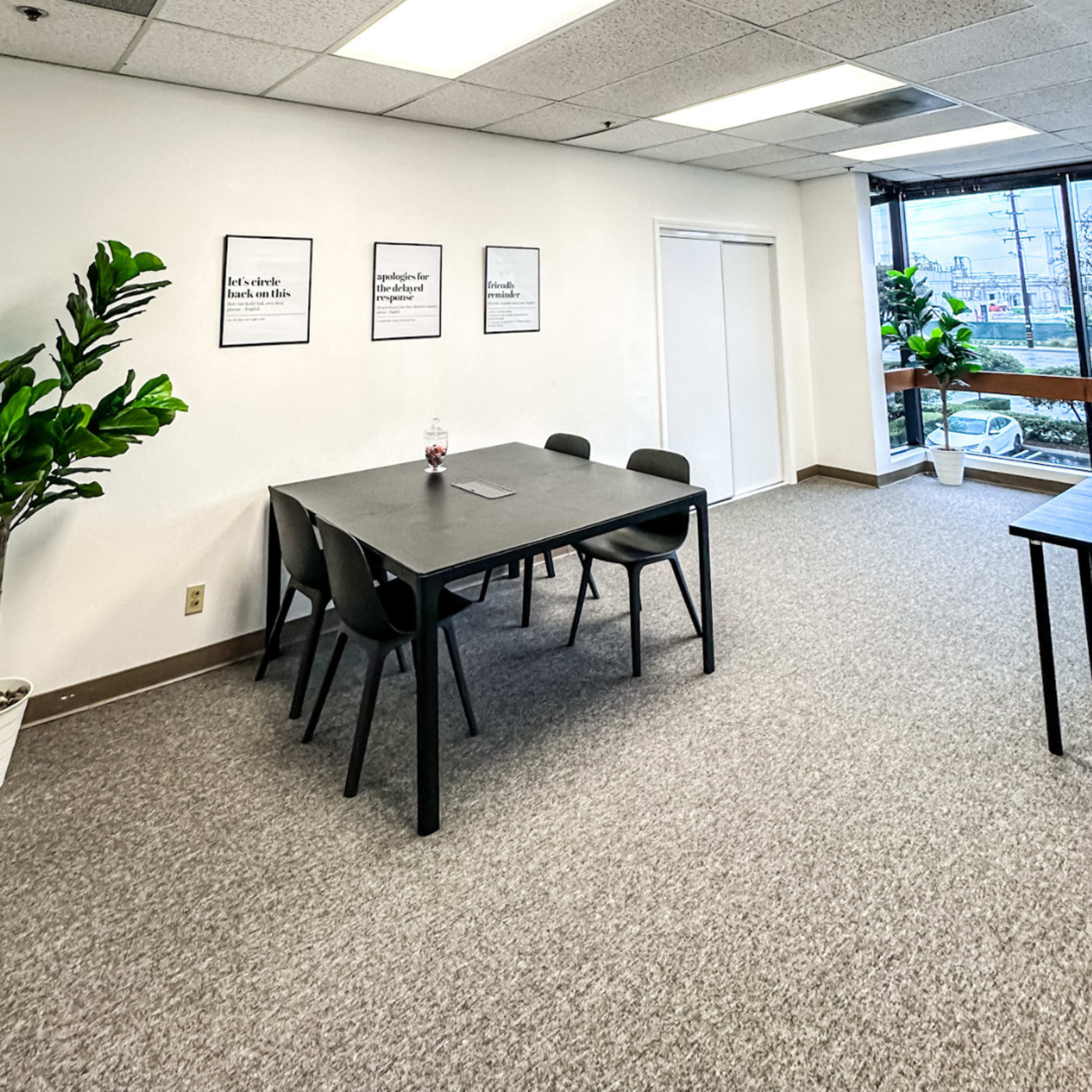 A modern office space features a large black table surrounded by chairs, with potted plants and framed quotes on the walls.