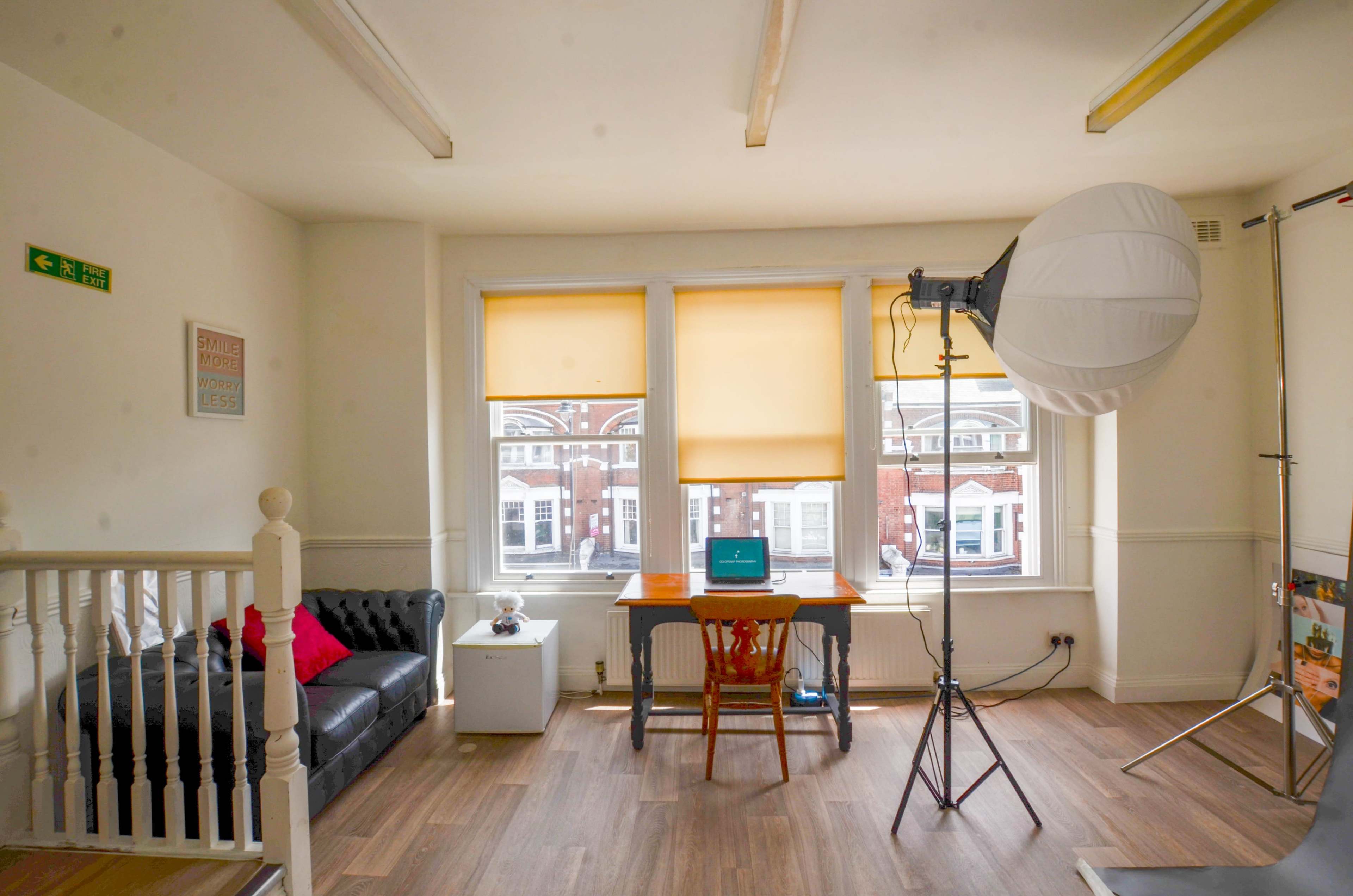 The image shows a bright room with large windows featuring yellow blinds, a wooden table with a laptop, a chair, a black sofa, and studio lighting equipment.