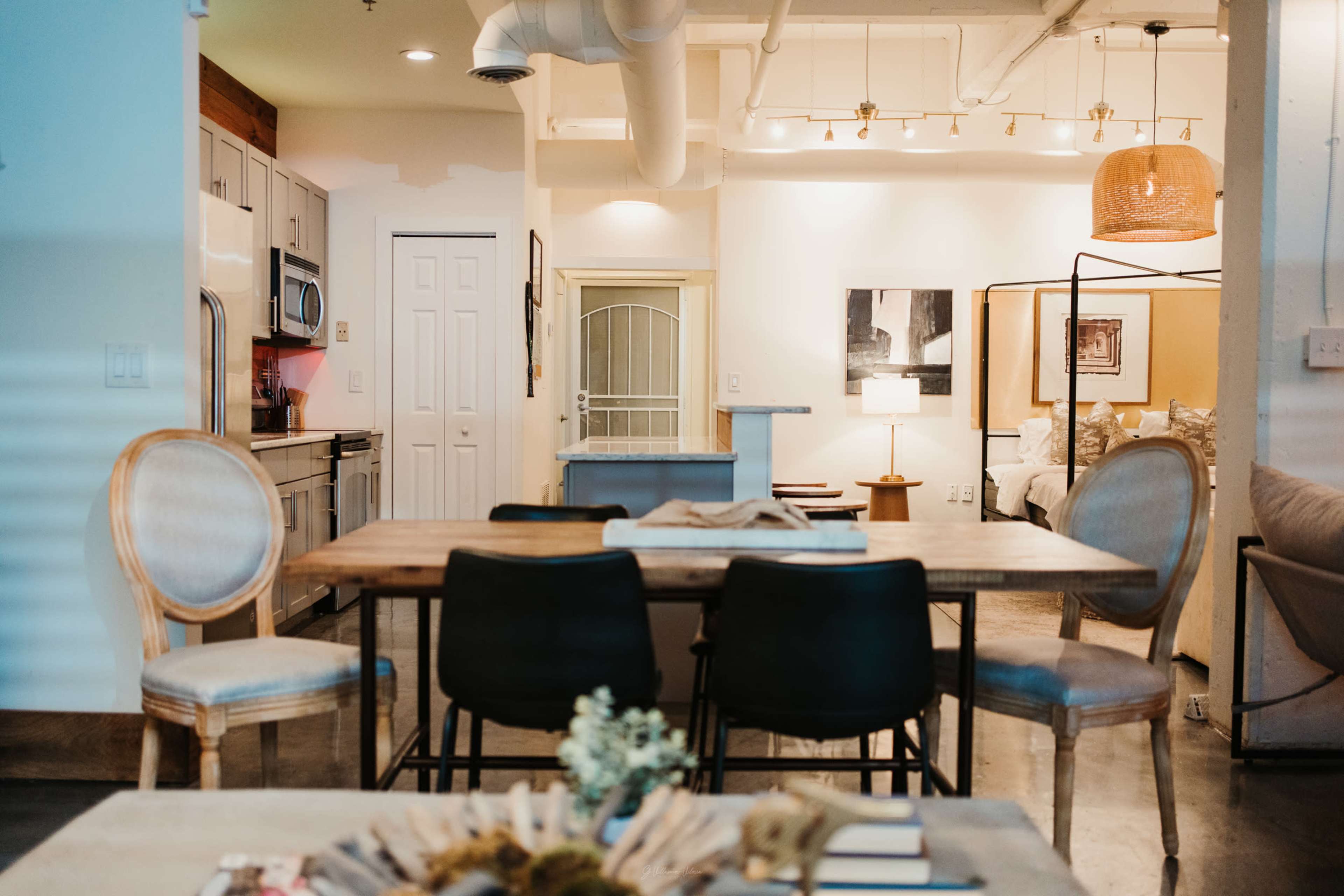 The image shows a modern open-concept living space featuring a dining table with chairs, a kitchen area, and a bedroom visible in the background.