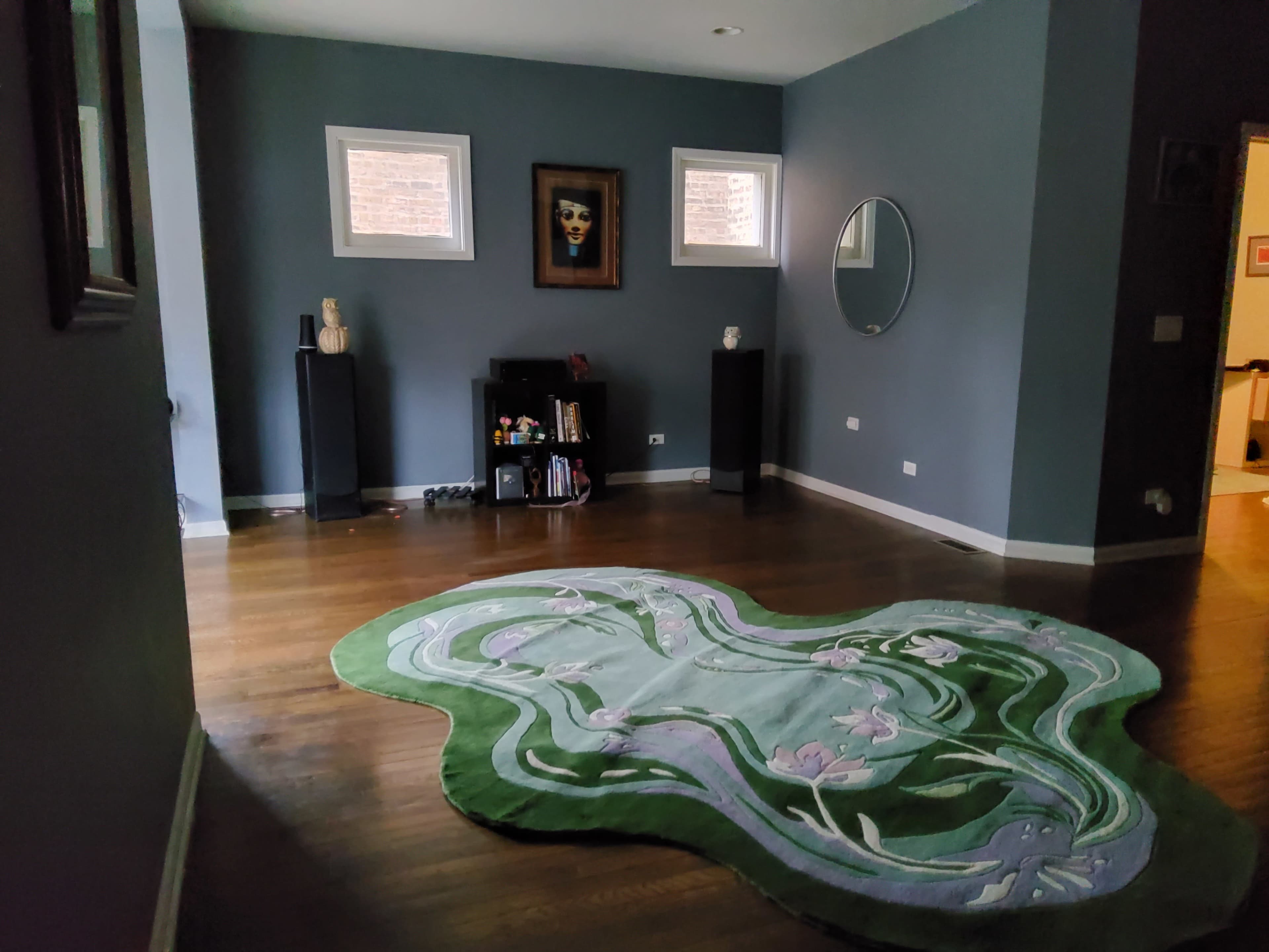 The room features hardwood floors, a large green area rug with a floral design, a black bookshelf, and a round mirror on the wall.