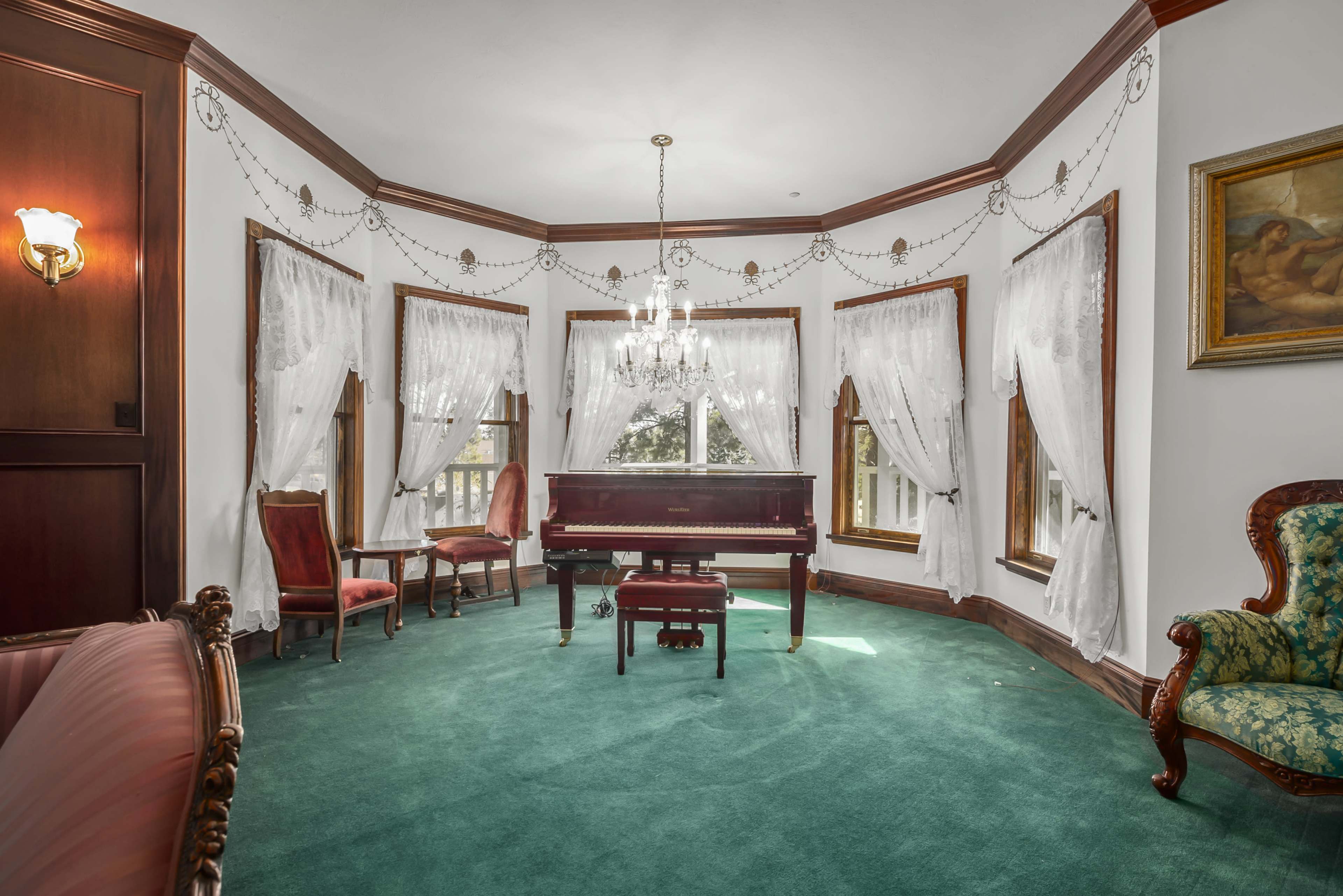 The room features a grand piano centered on a green carpet, surrounded by elegant seating and large windows adorned with draped curtains.