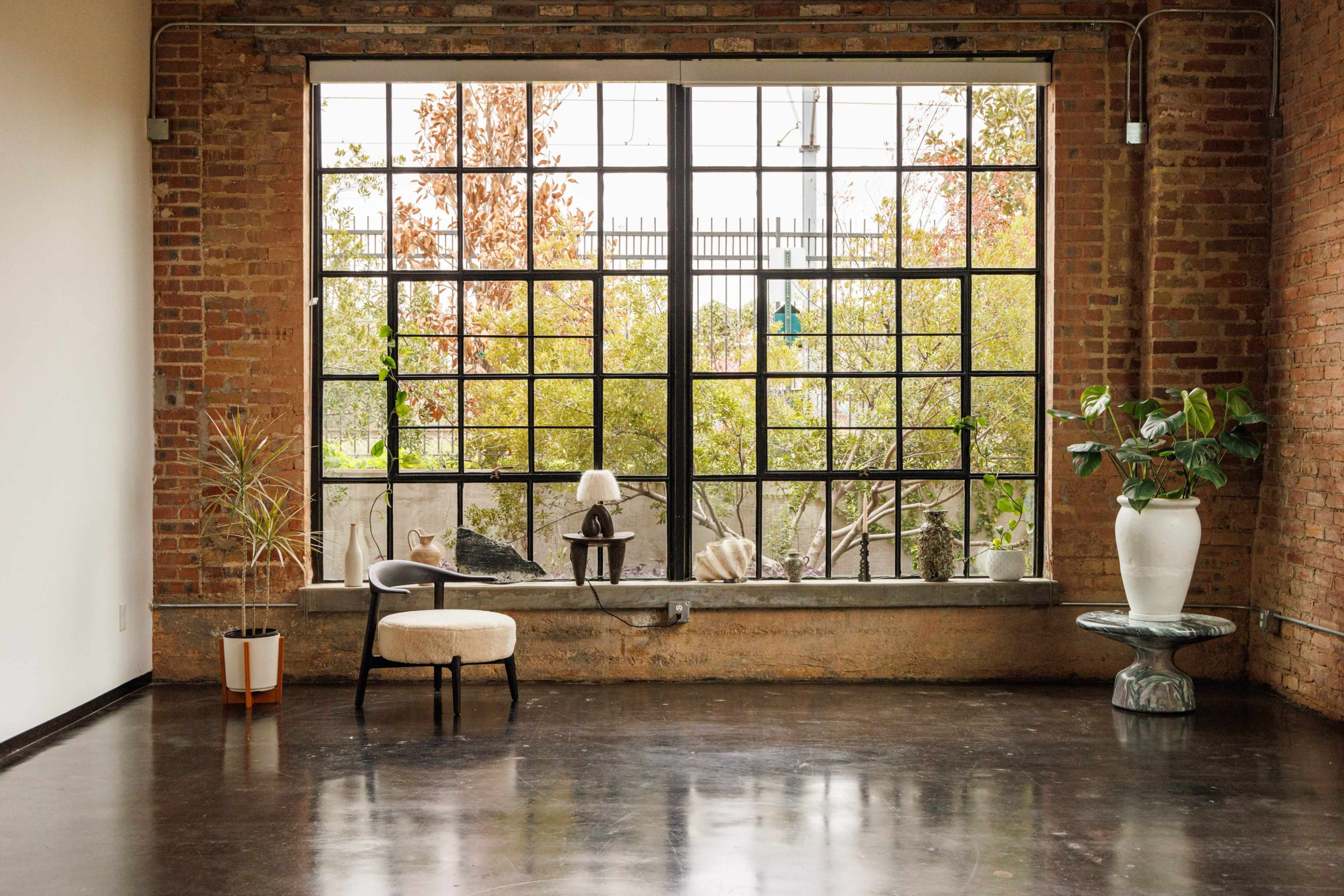 The image shows a spacious room with large industrial-style windows, exposing a view of greenery outside, and minimalistic furniture including a chair, lamp, and potted plants.