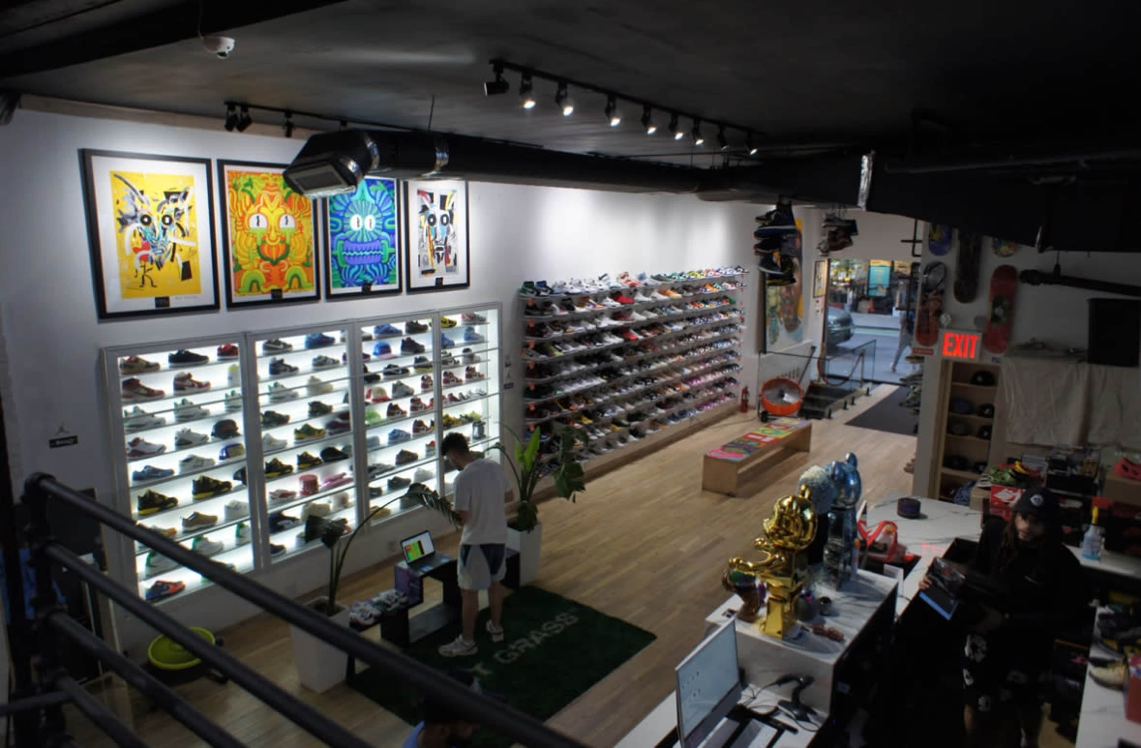 A retail store featuring a wall of colorful sneakers, artwork on the walls, and a customer browsing near the checkout area.