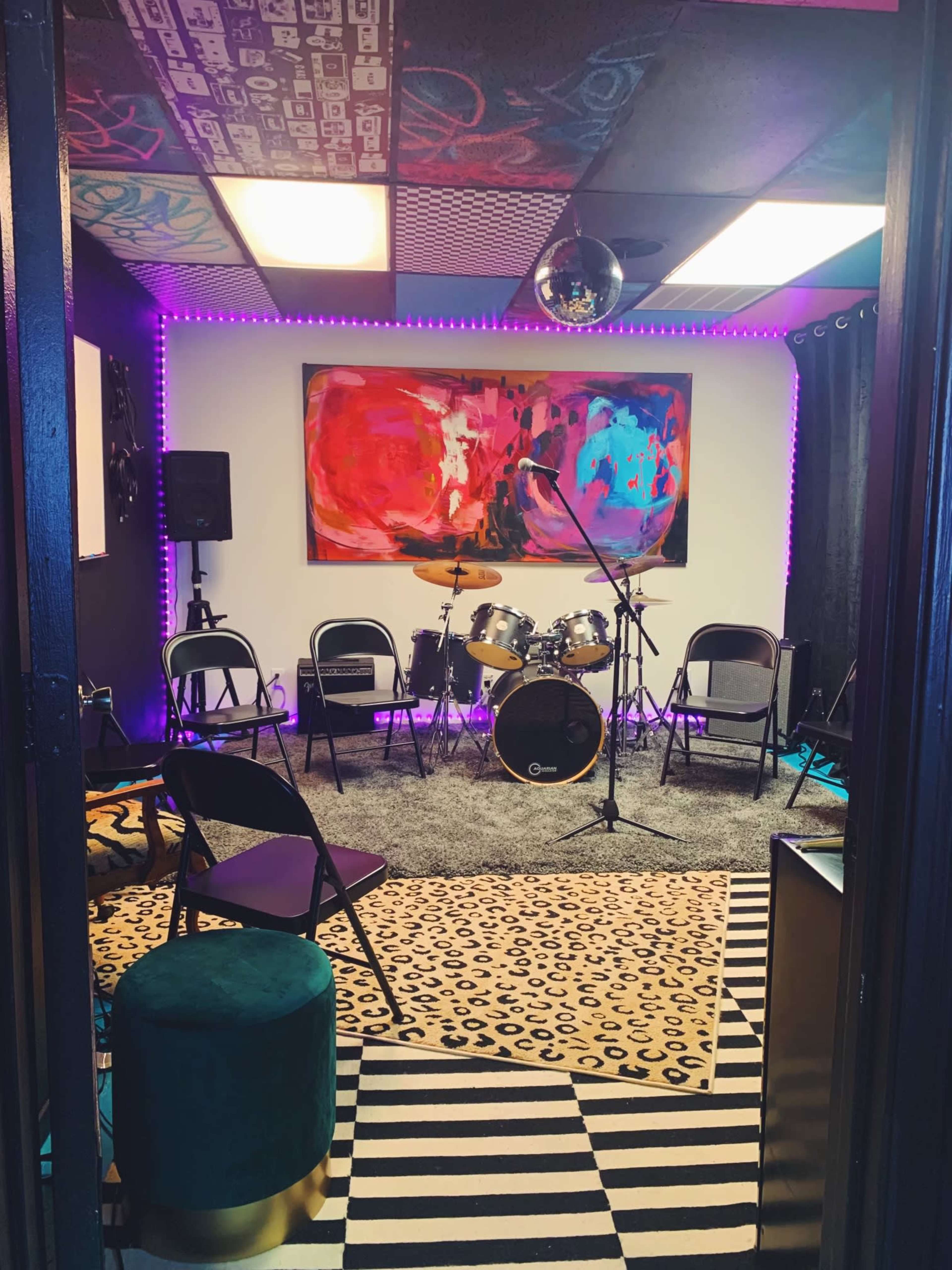 A music rehearsal space features a drum set, chairs, and colorful artwork, illuminated by purple LED lights.