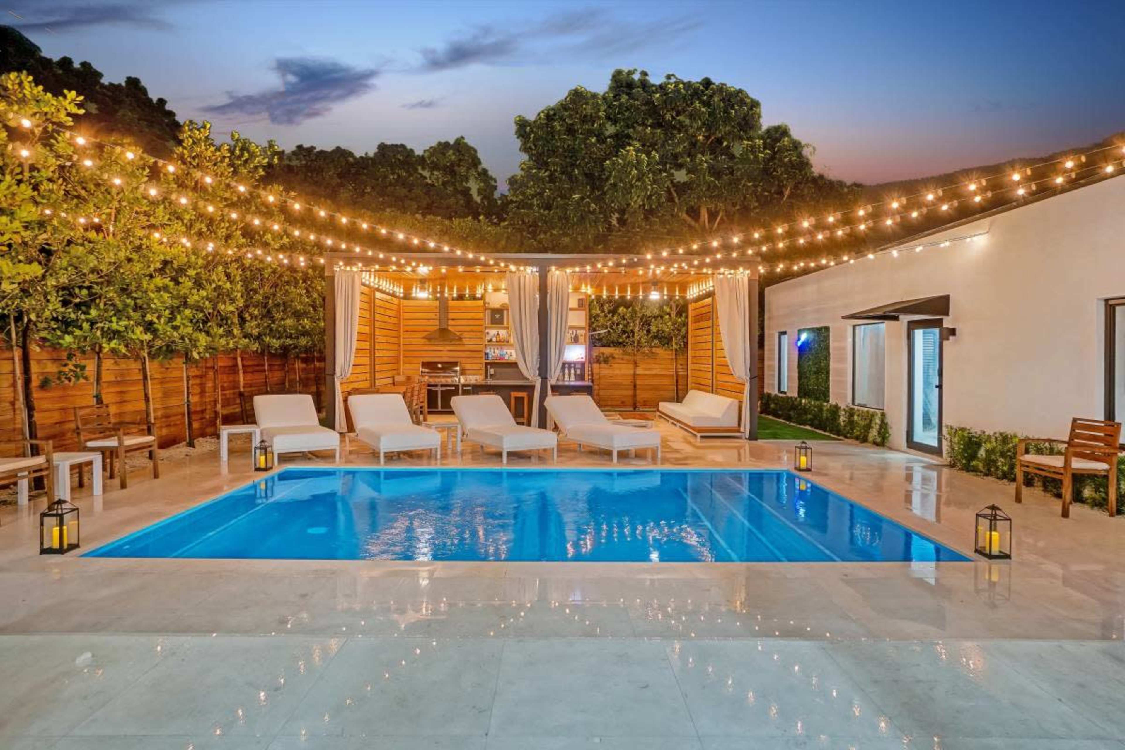 The image shows a well-lit backyard with a swimming pool surrounded by lounge chairs, decorative lanterns, and string lights, set against a twilight sky.
