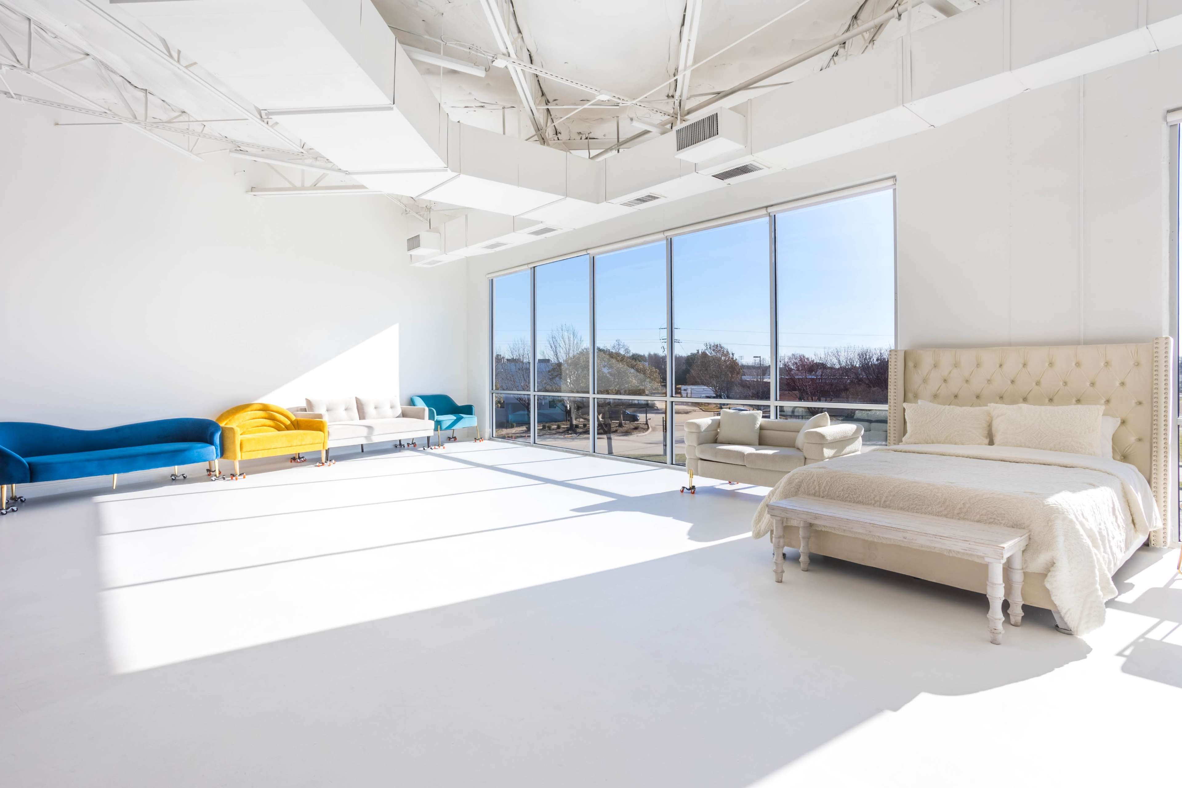 A bright, spacious room features a bed and several couches near large windows, with white walls and a light-colored floor.