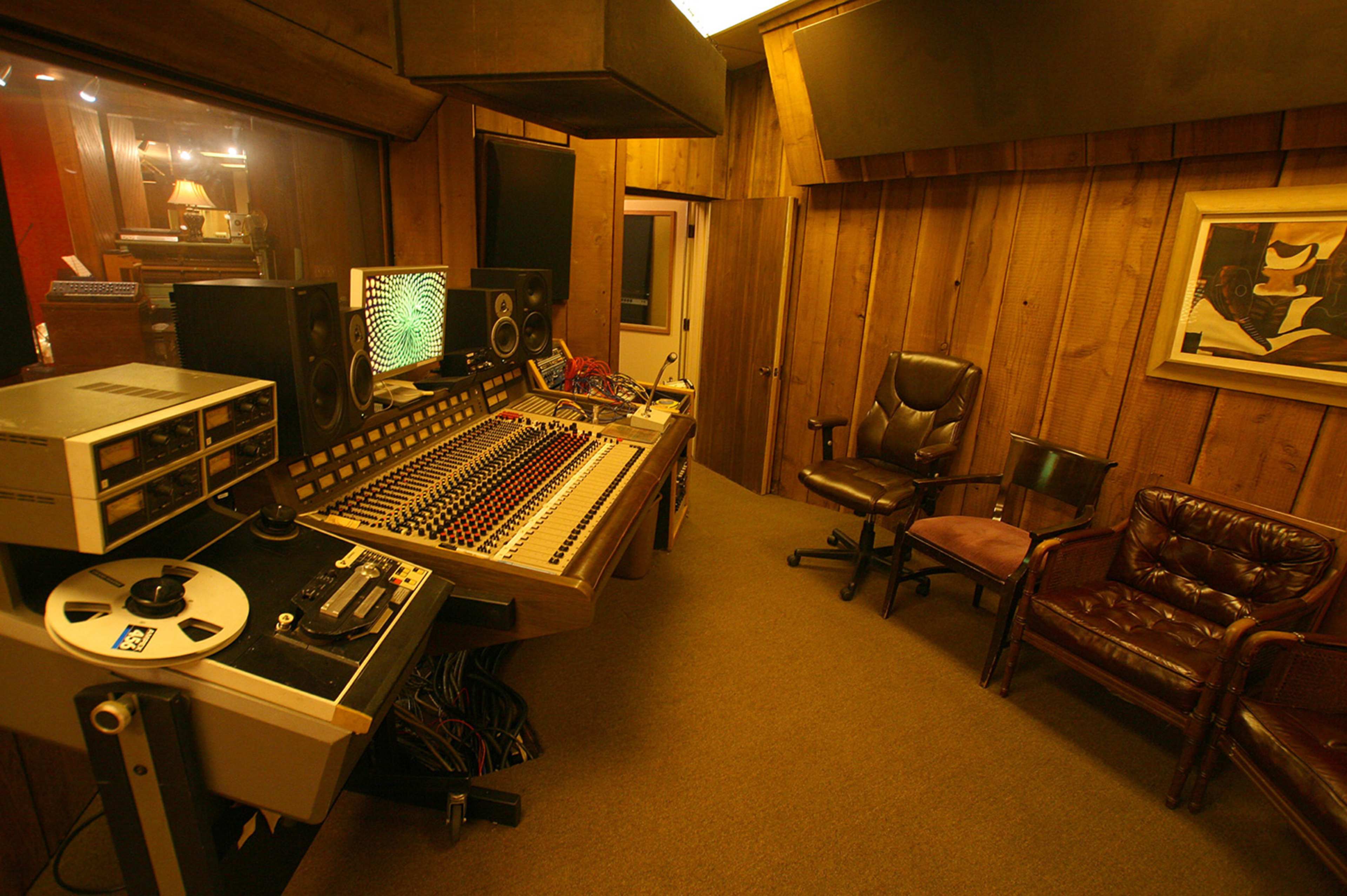A recording studio with a mixing console, tape machine, and two chairs placed in a wooden-paneled room.