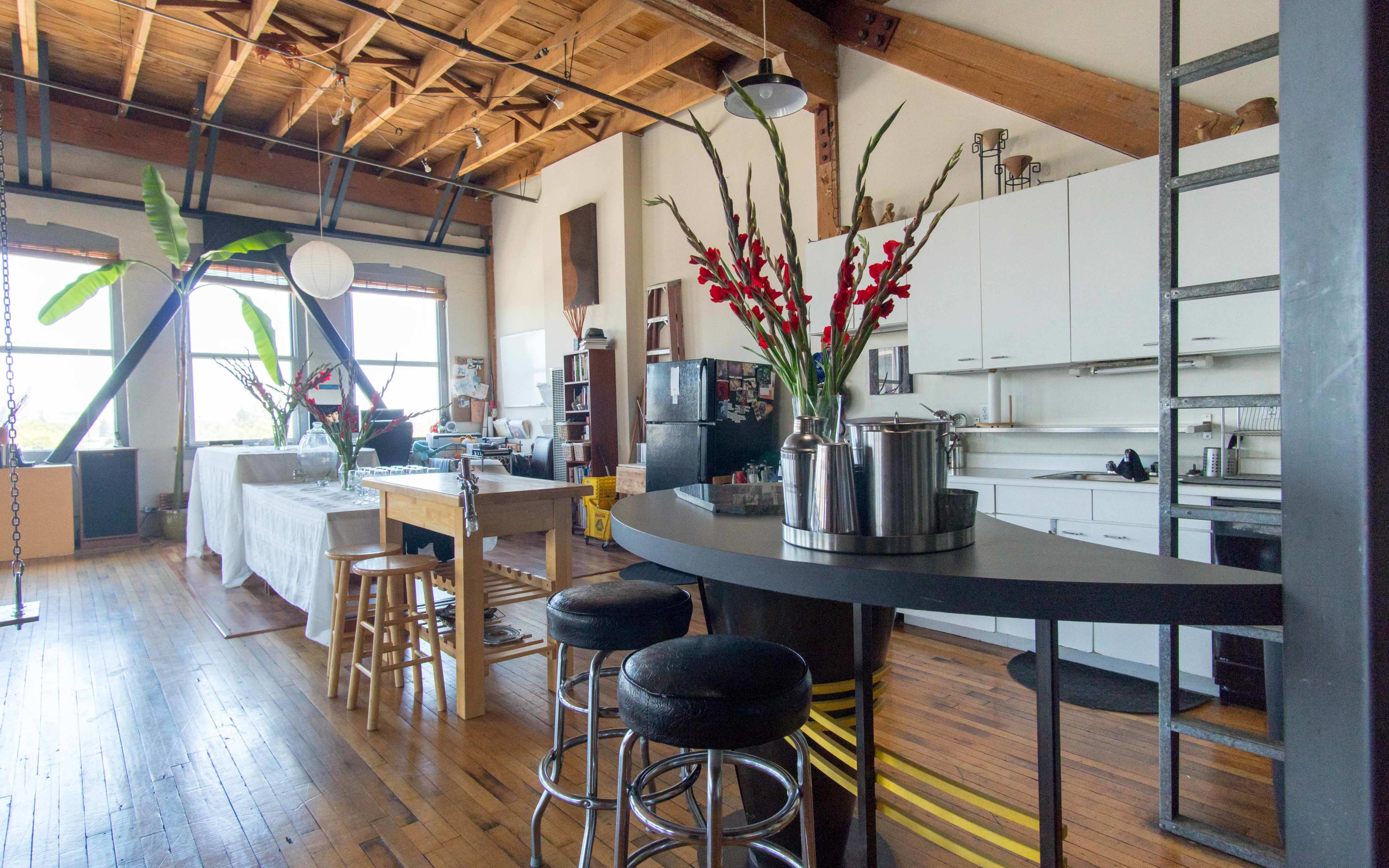 The image shows a spacious, airy loft-style kitchen and dining area with wooden floors, a central island, and large windows letting in natural light.