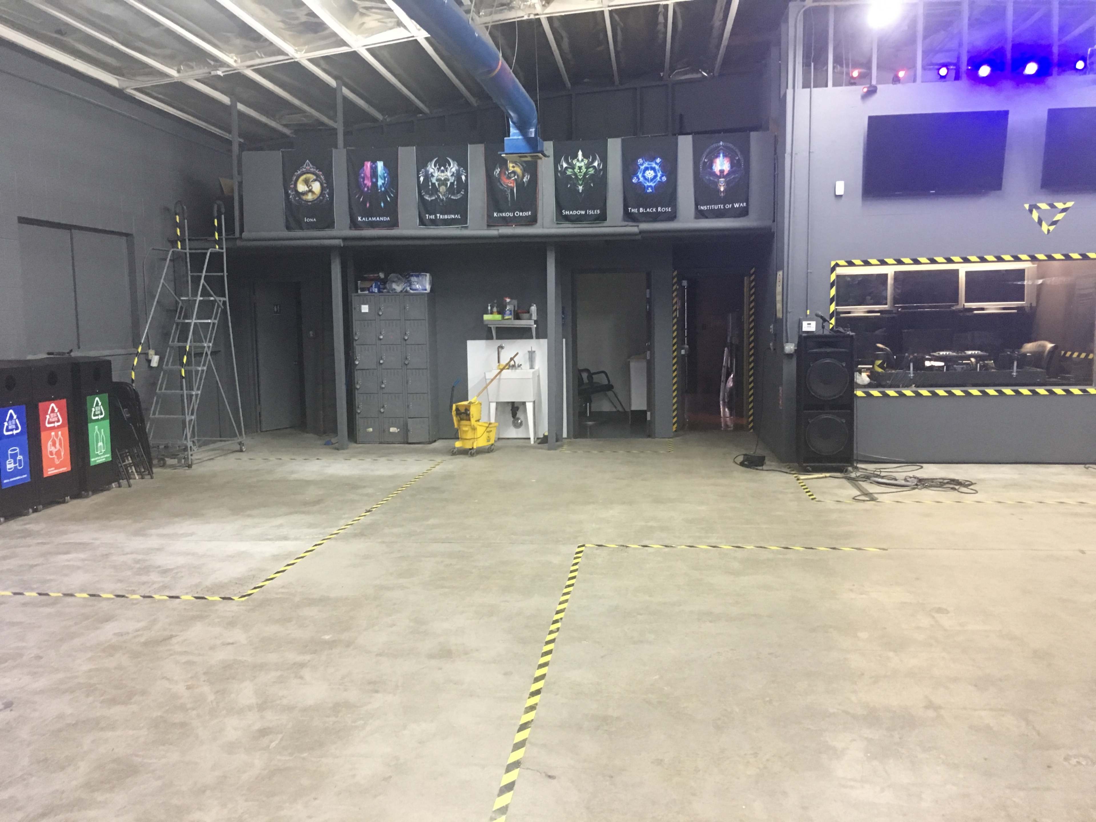 An industrial-style room with a concrete floor, featuring lockers, posters on the walls, and a cleaning cart in the center.