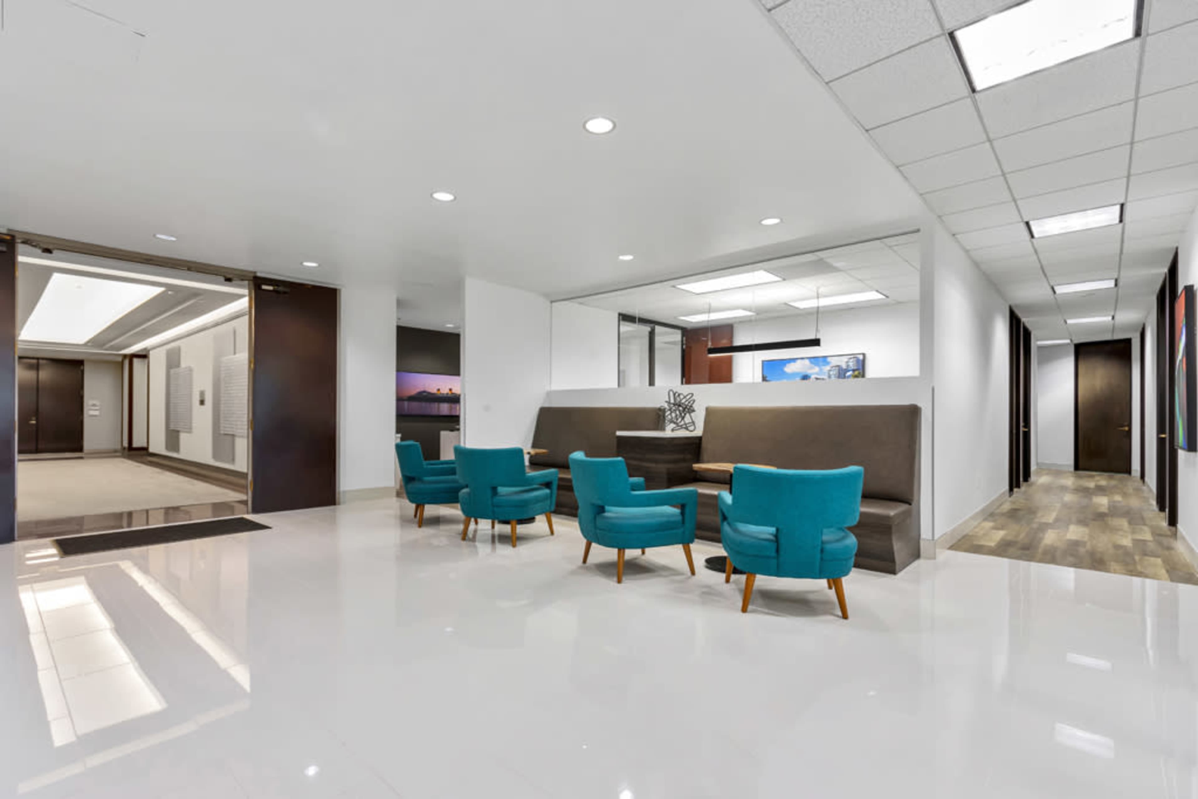 The image shows a modern office lobby with teal chairs arranged around a low table, white walls, and a corridor leading to additional office spaces.