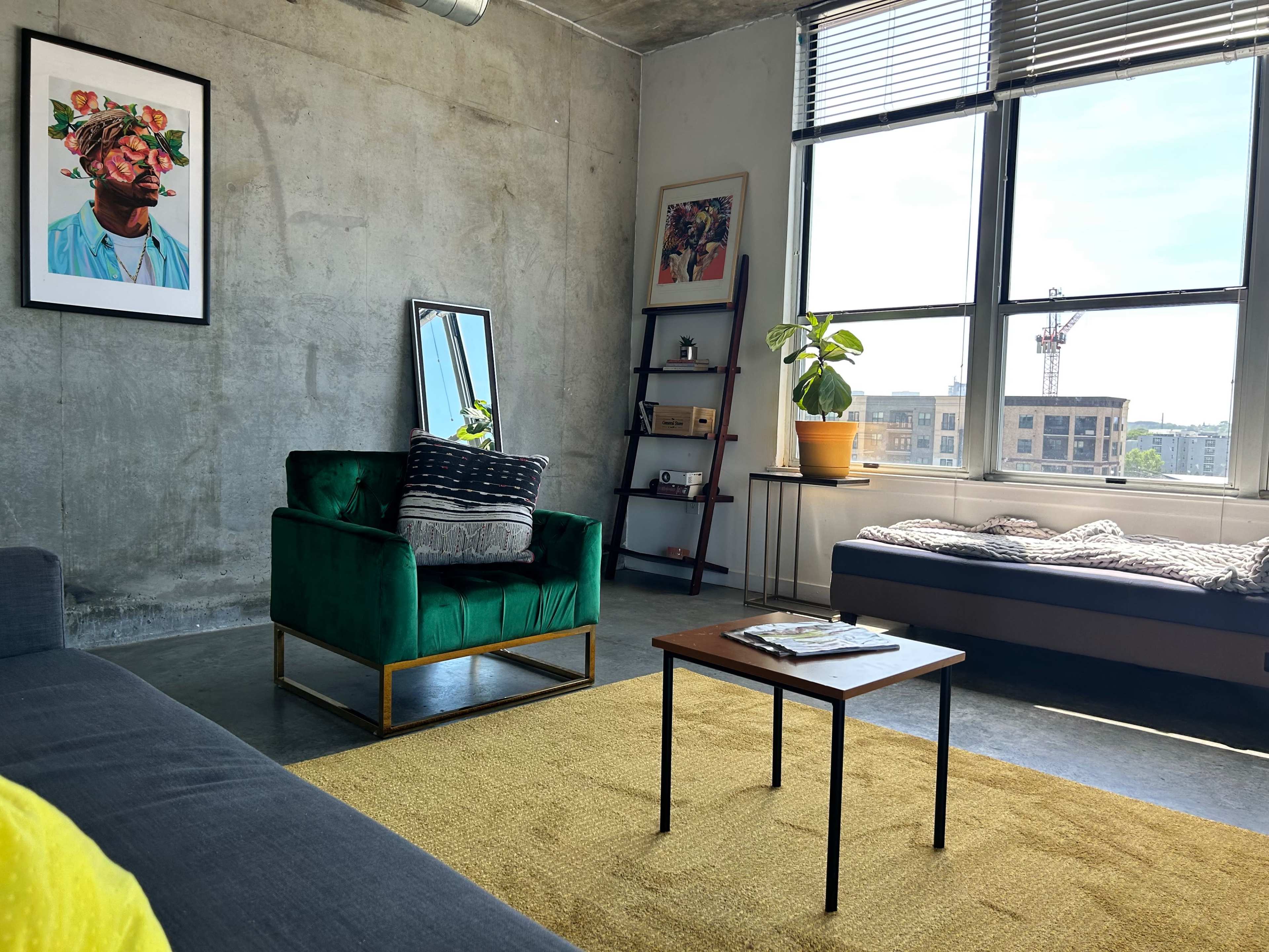 The image shows a modern living room featuring a green velvet chair, a wooden coffee table, a large window with natural light, and a mix of artwork on the walls.