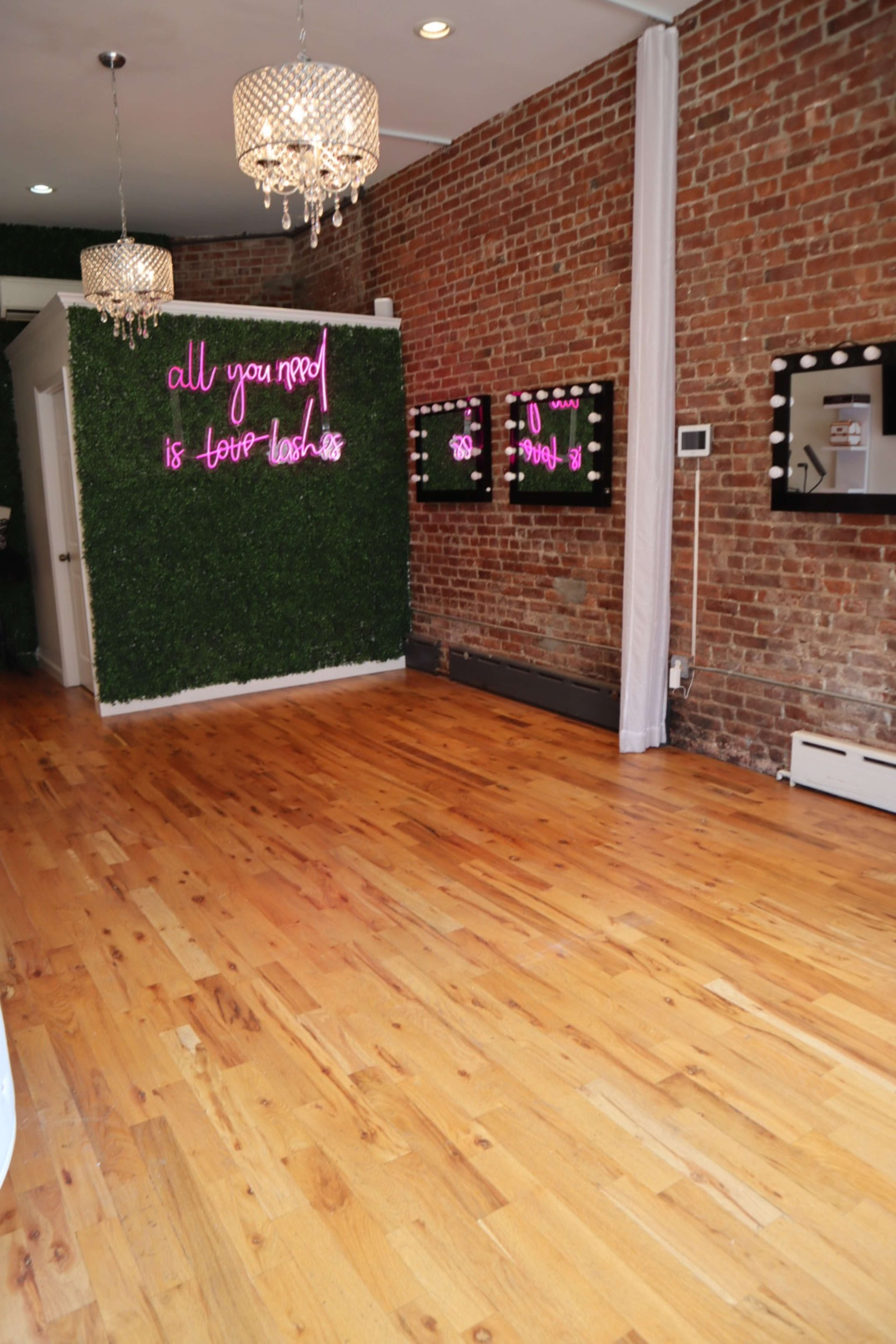 The image shows a stylish room with exposed brick walls, wooden flooring, and neon signs, featuring decorative mirrors framed with light bulbs.