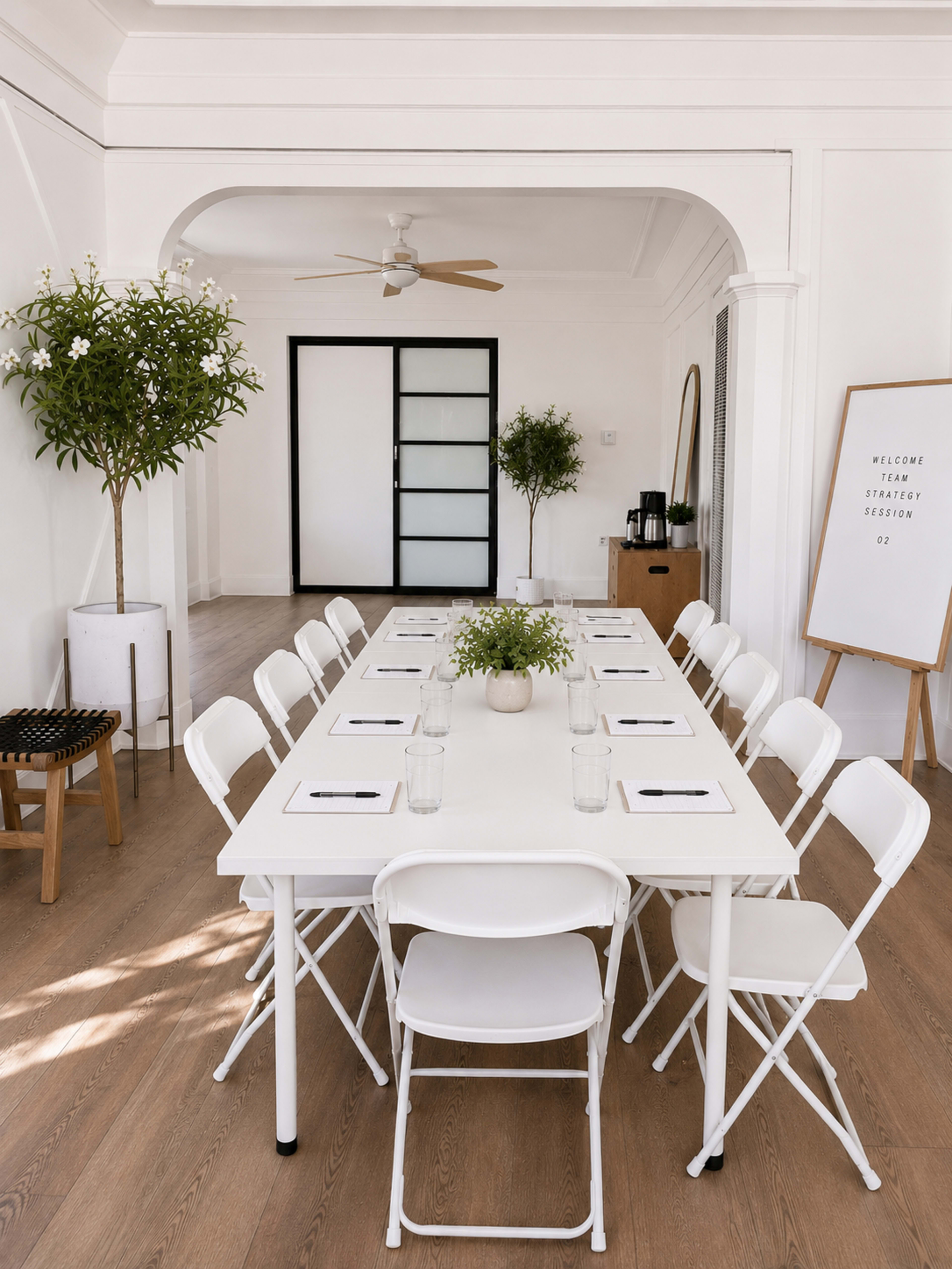A long white table with chairs surrounds it in a bright, minimalistic meeting room featuring potted plants and a whiteboard.