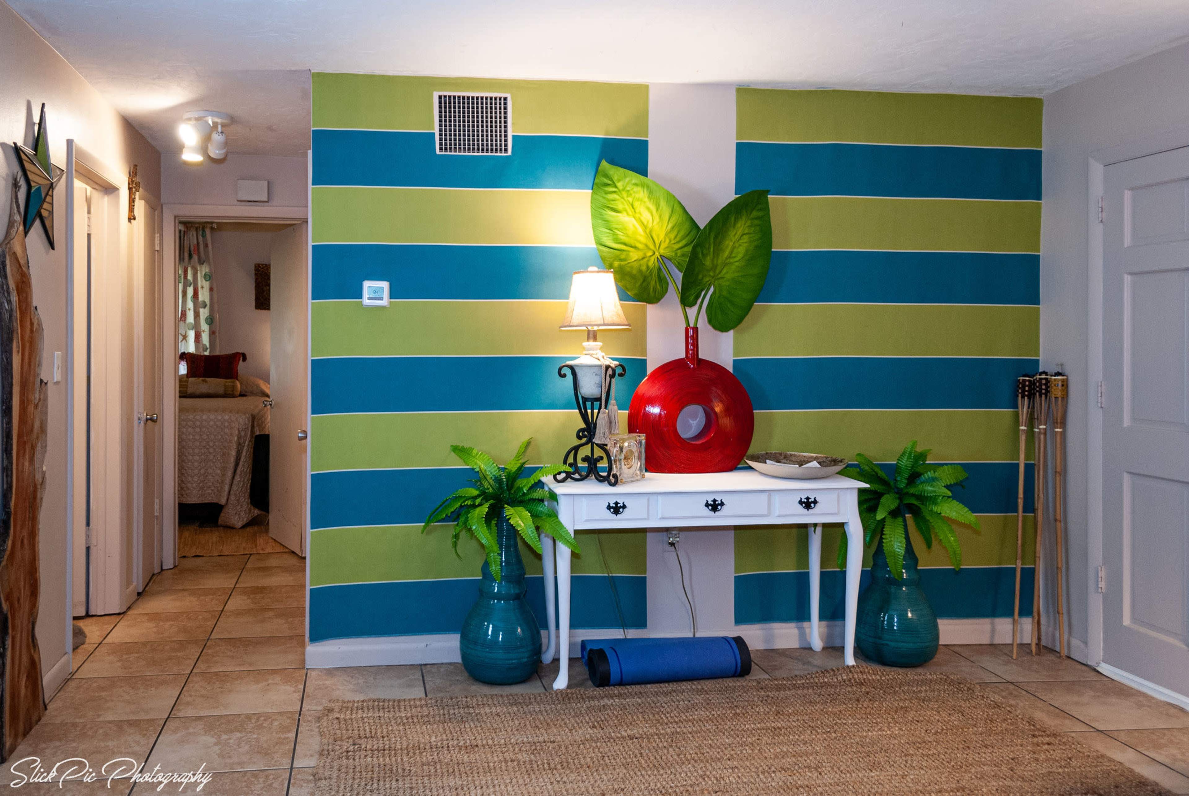 The image shows a brightly colored entryway featuring a green and blue striped wall, a white console table with a lamp, decorative plants, and a red circular artwork.