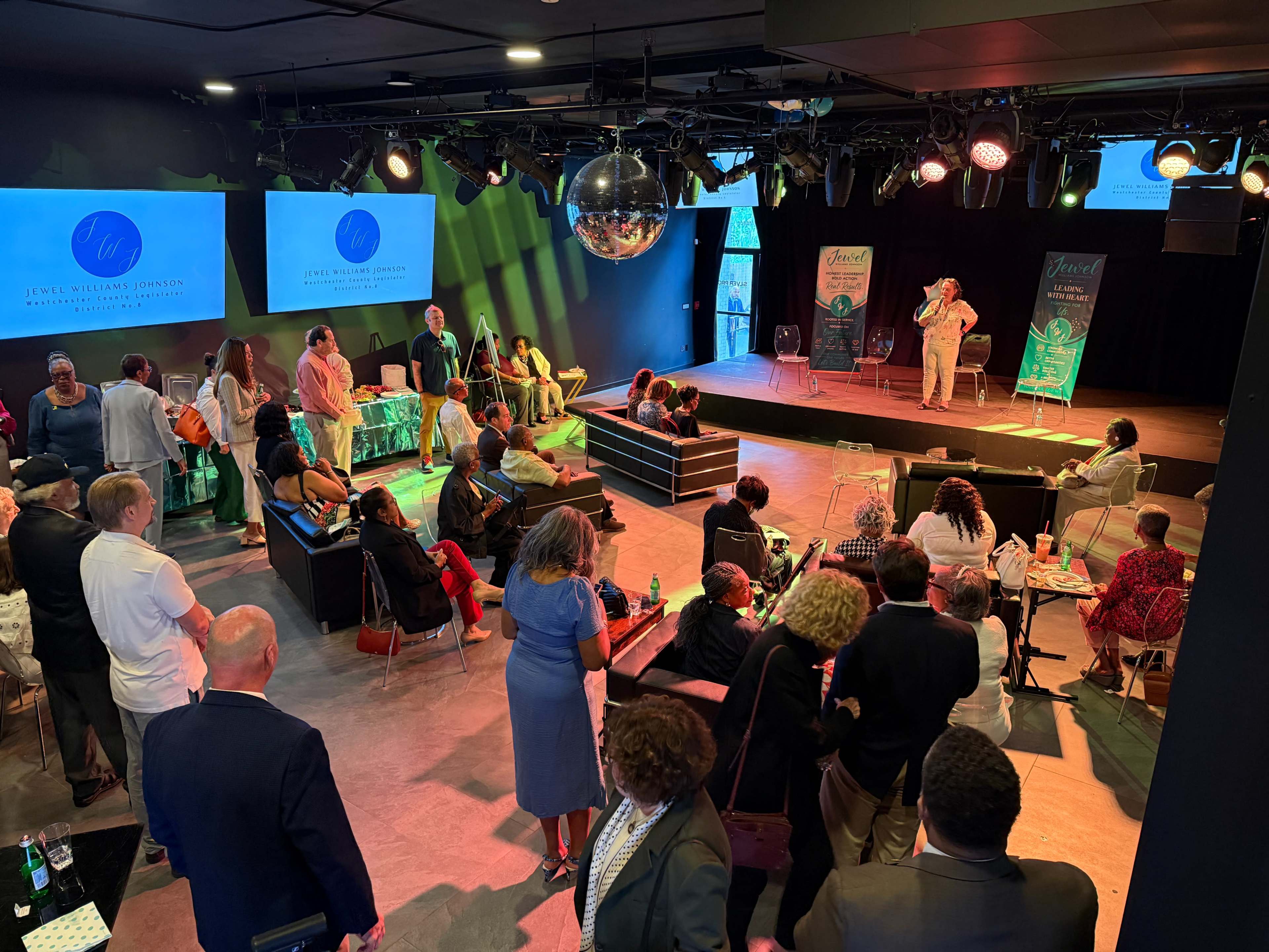 A crowd gathers in a modern event space featuring a stage with lighting, a disco ball overhead, and screens displaying promotional material.
