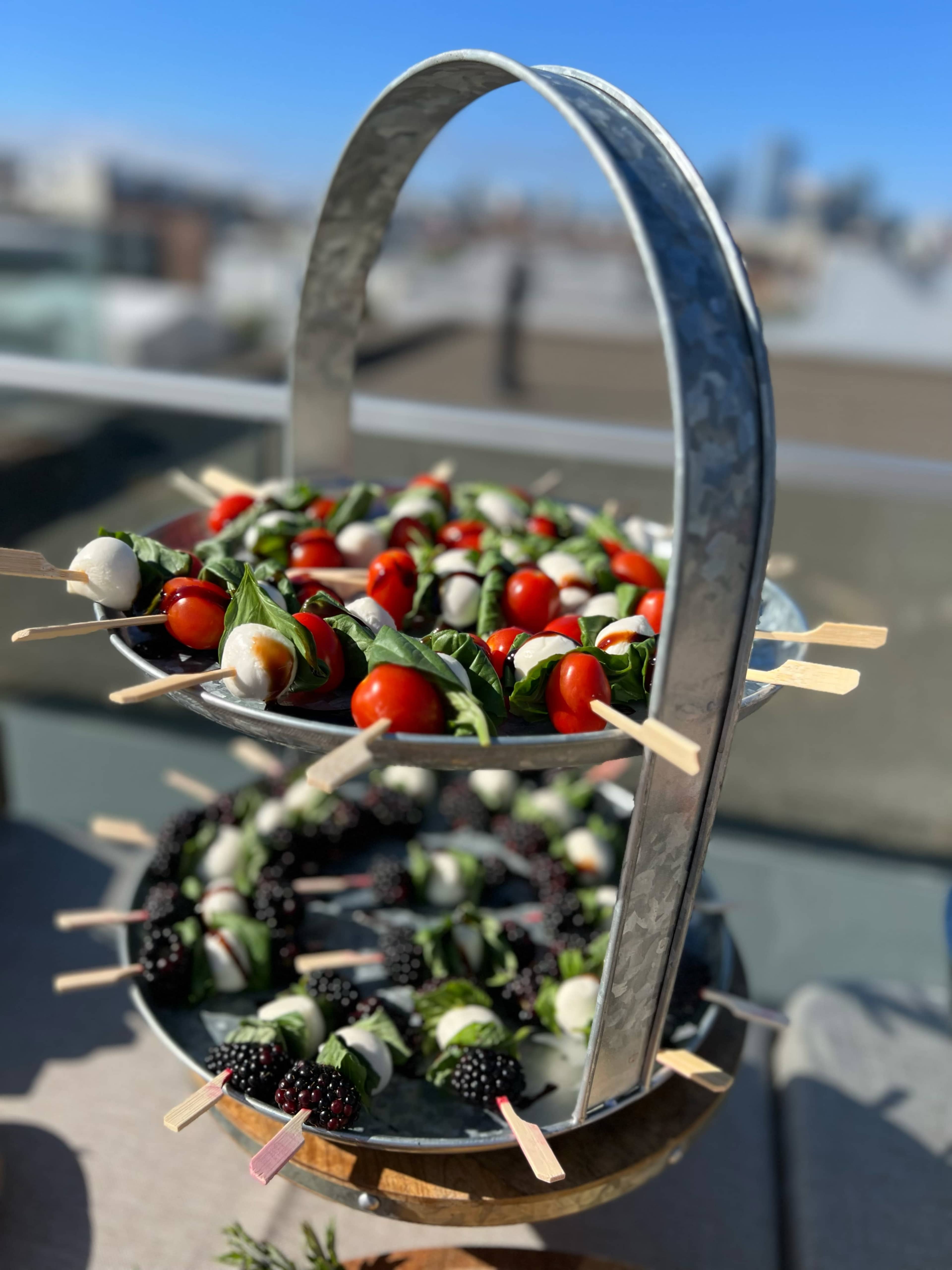A two-tiered serving tray displays skewers of cherry tomatoes, basil, and mozzarella balls alongside blackberries.