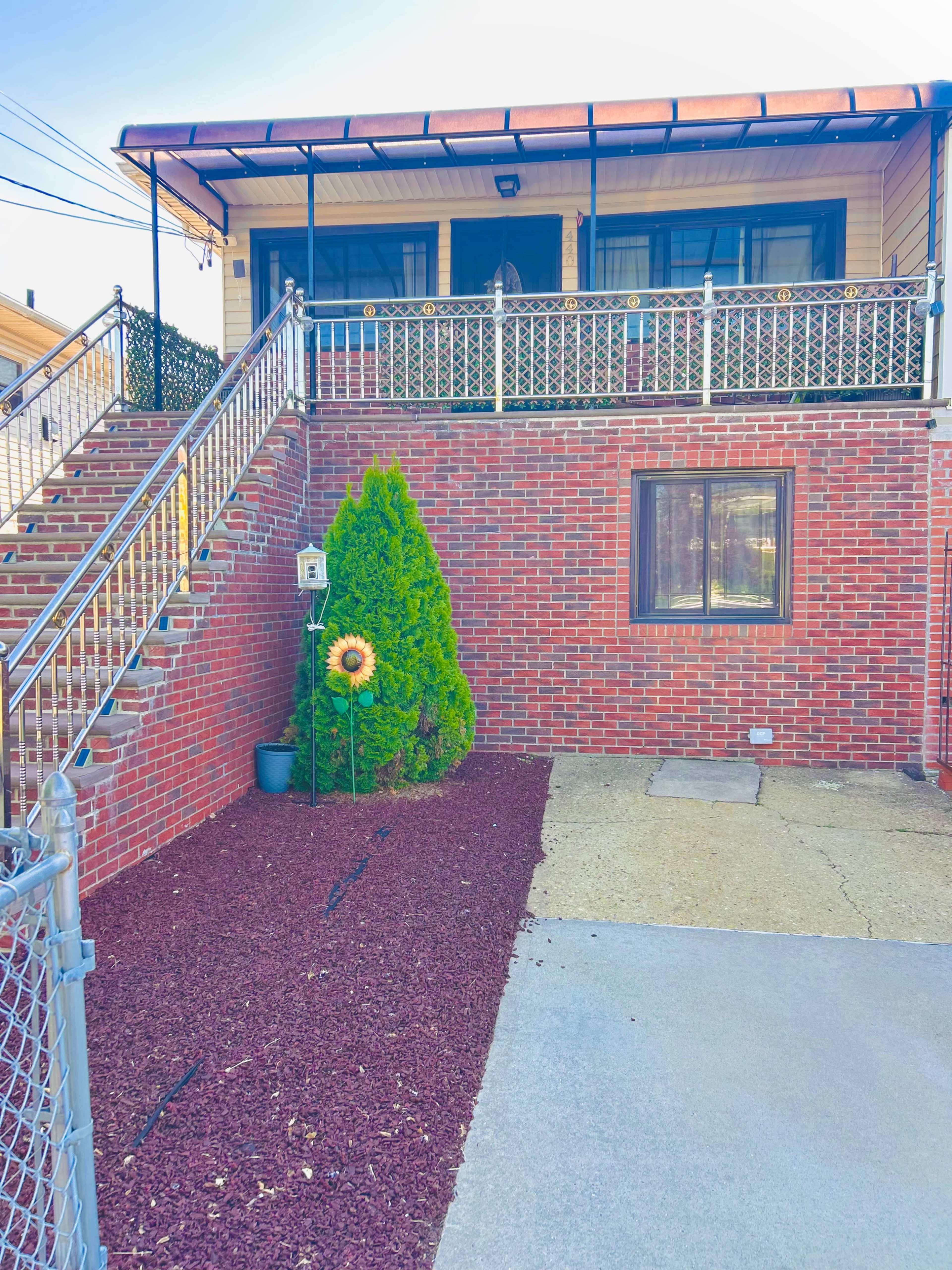 The image shows a brick house with a front staircase leading up to a porch, surrounded by a small yard featuring mulch and a sunflower decoration.