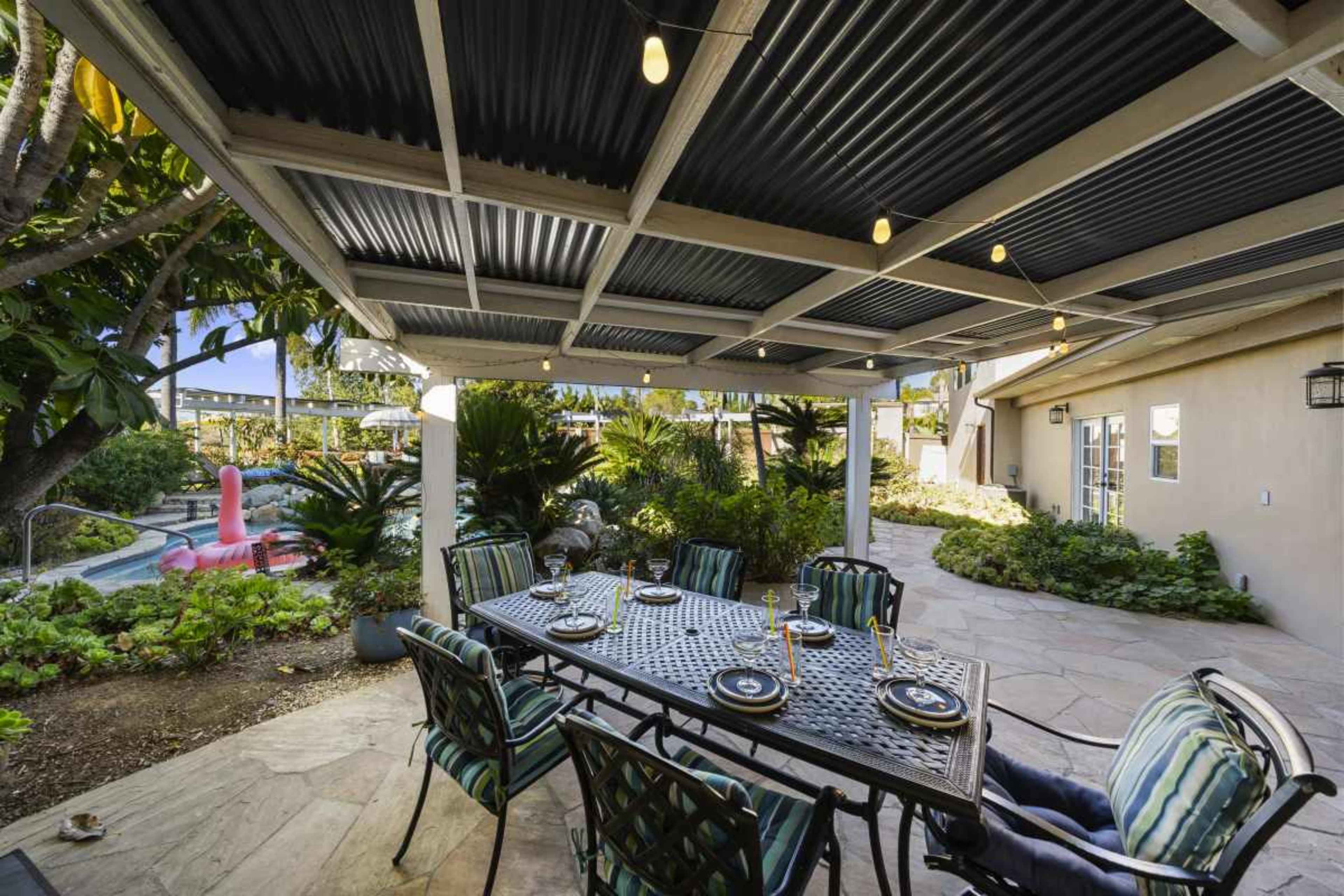 The image shows a covered outdoor dining area with a table set for a meal, surrounded by lush greenery and a swimming pool in the background.