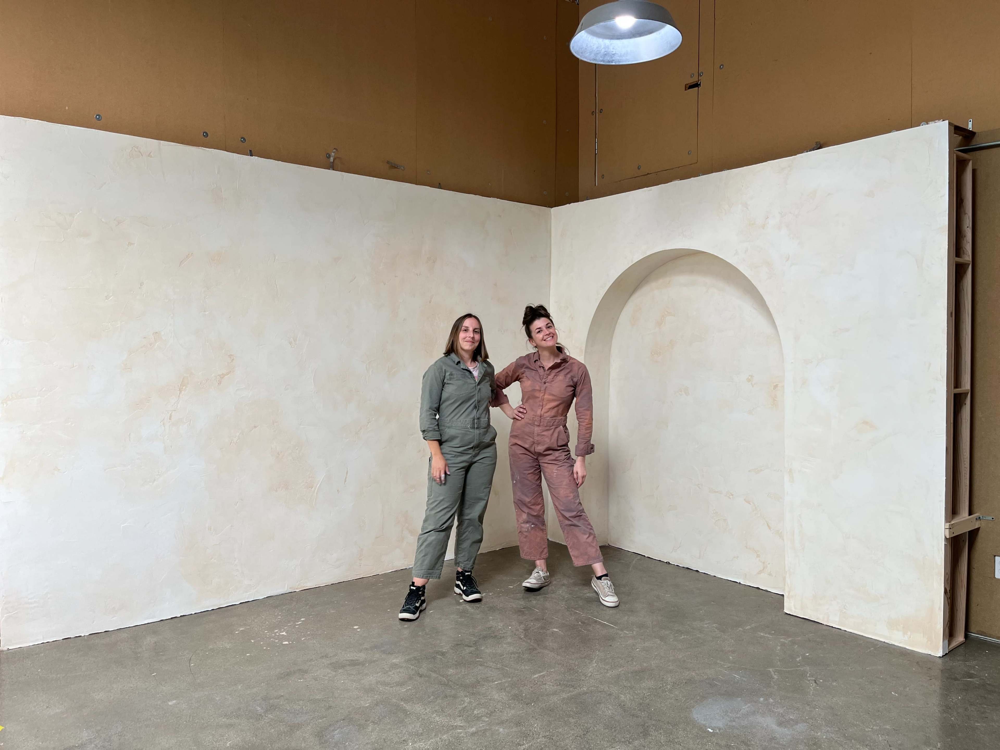 Two women stand in a minimalistic studio space with a large, arched opening in the wall behind them.