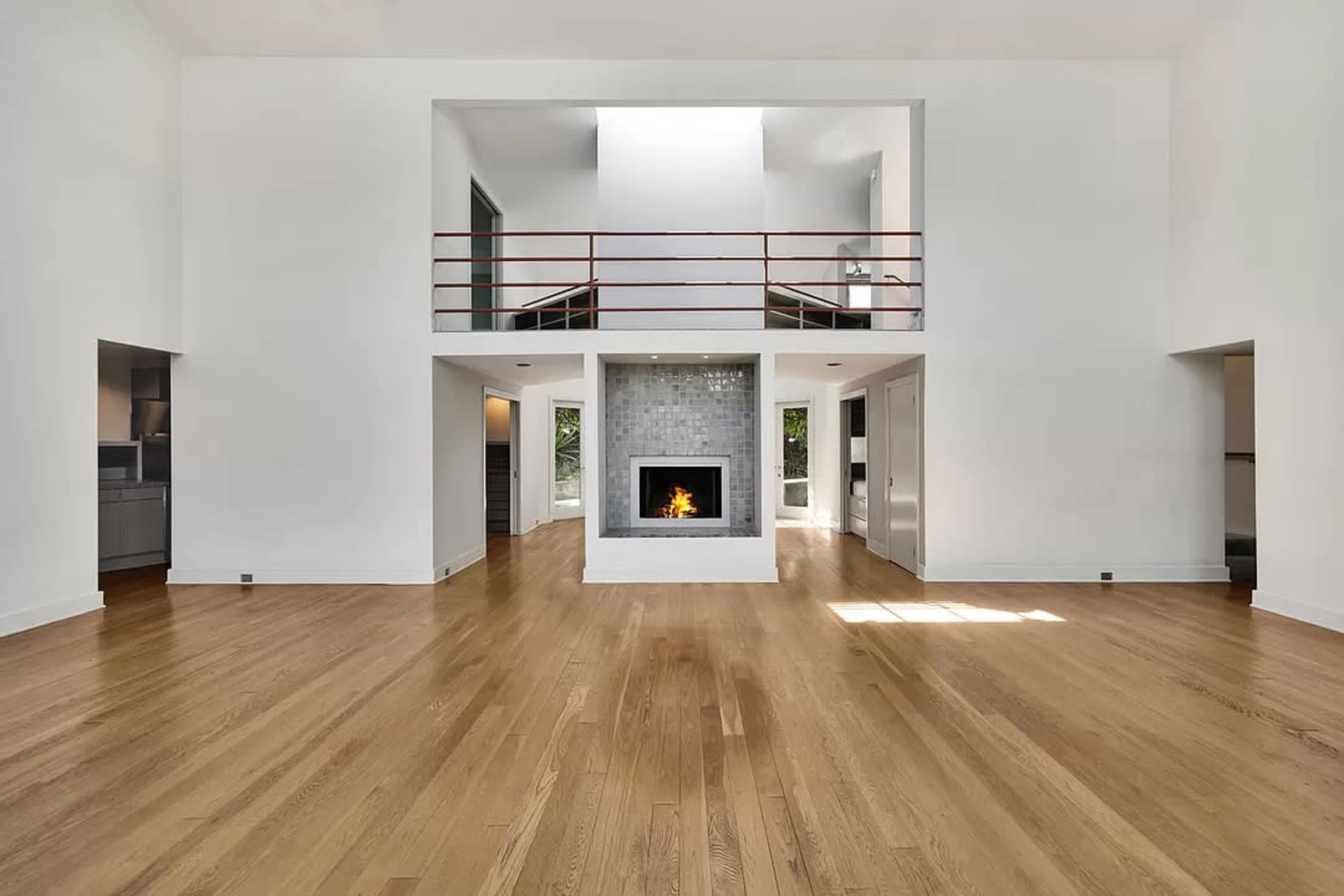 The image shows a modern living room with a central fireplace, high ceilings, and large windows allowing natural light to enter.