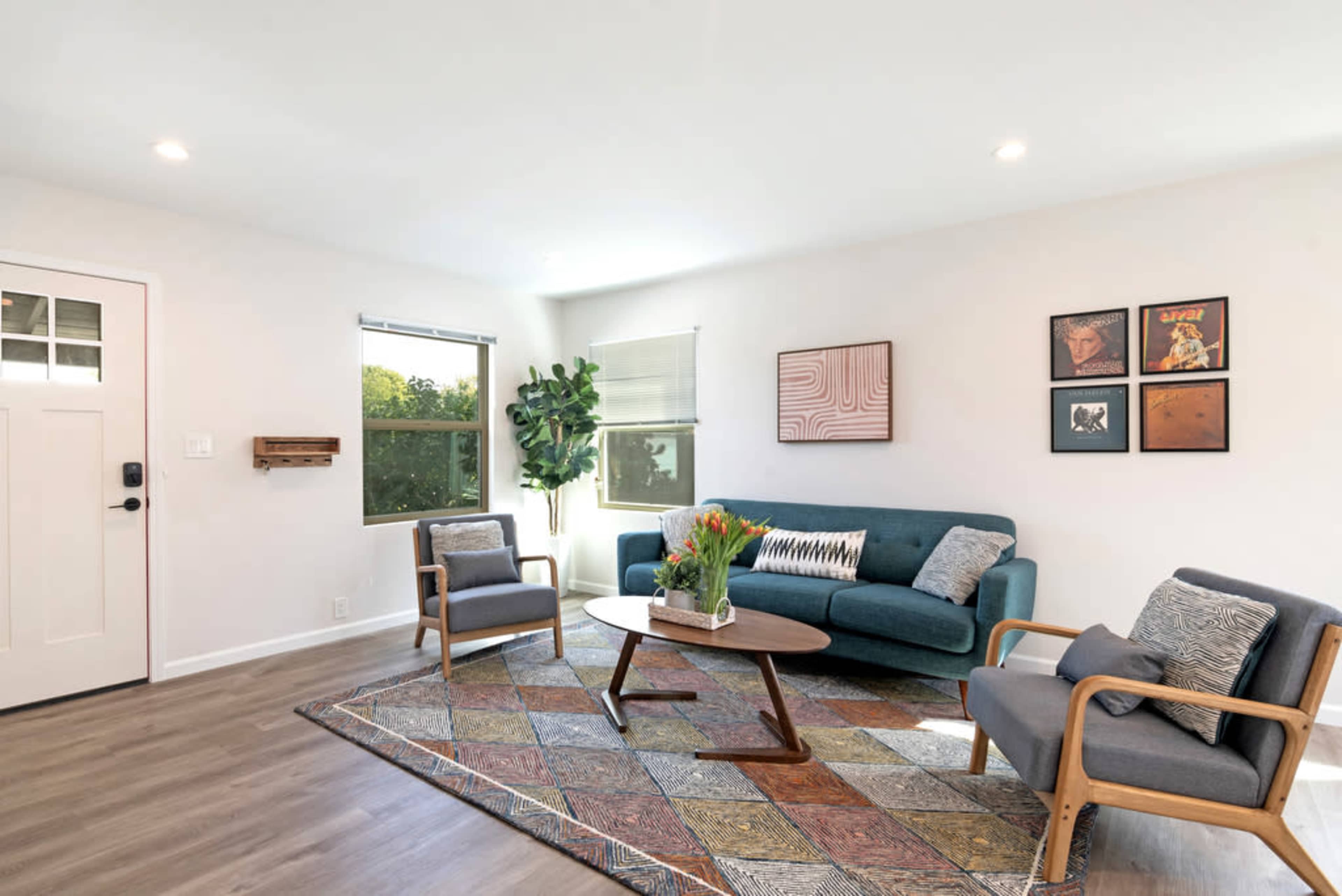 A modern living room features a green sofa, two wooden armchairs, a round coffee table, and a colorful patterned rug, with large windows allowing natural light.