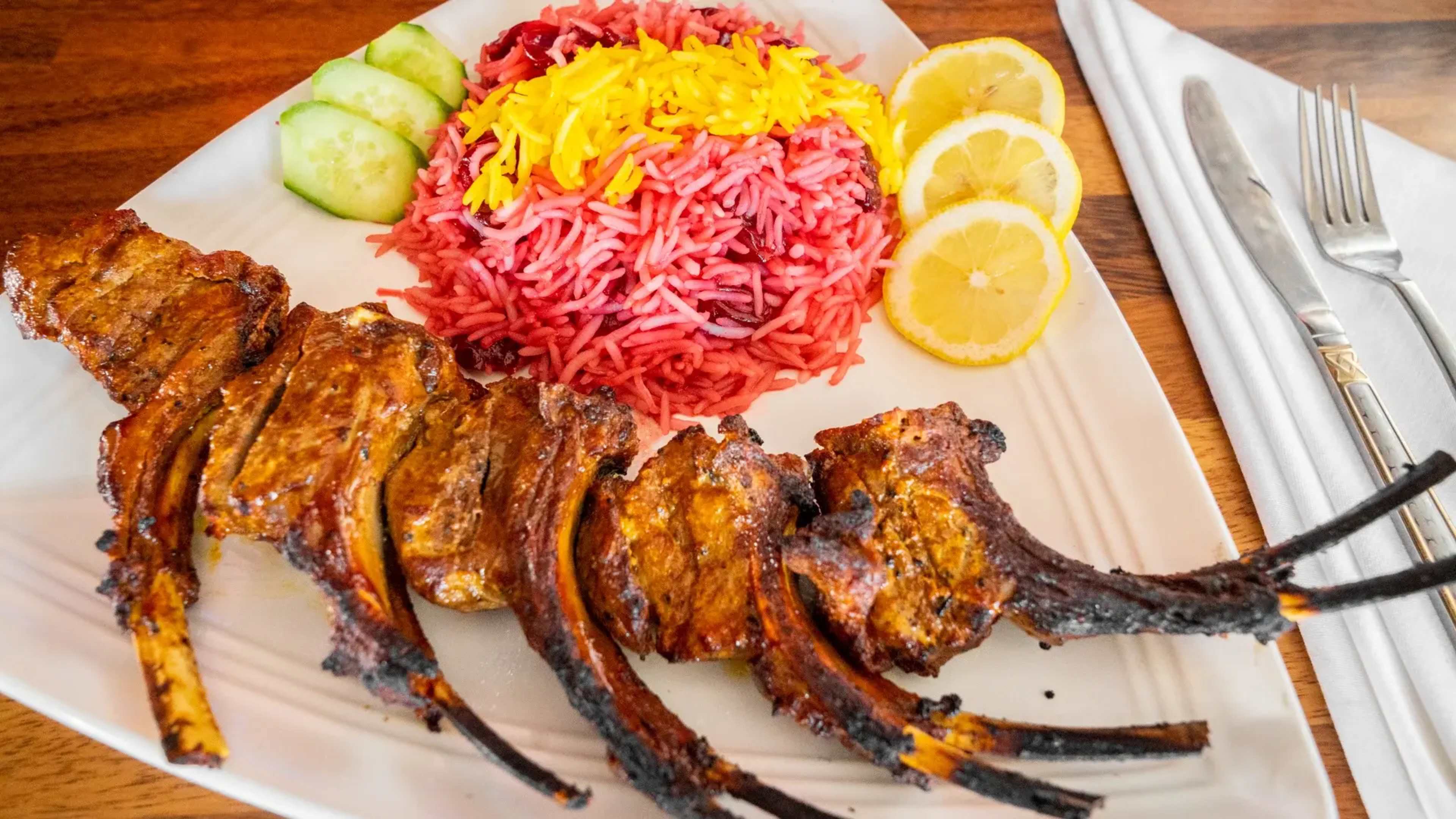 The dish features grilled lamb chops alongside pink rice garnished with yellow strands, accompanied by cucumber slices and lemon wedges.