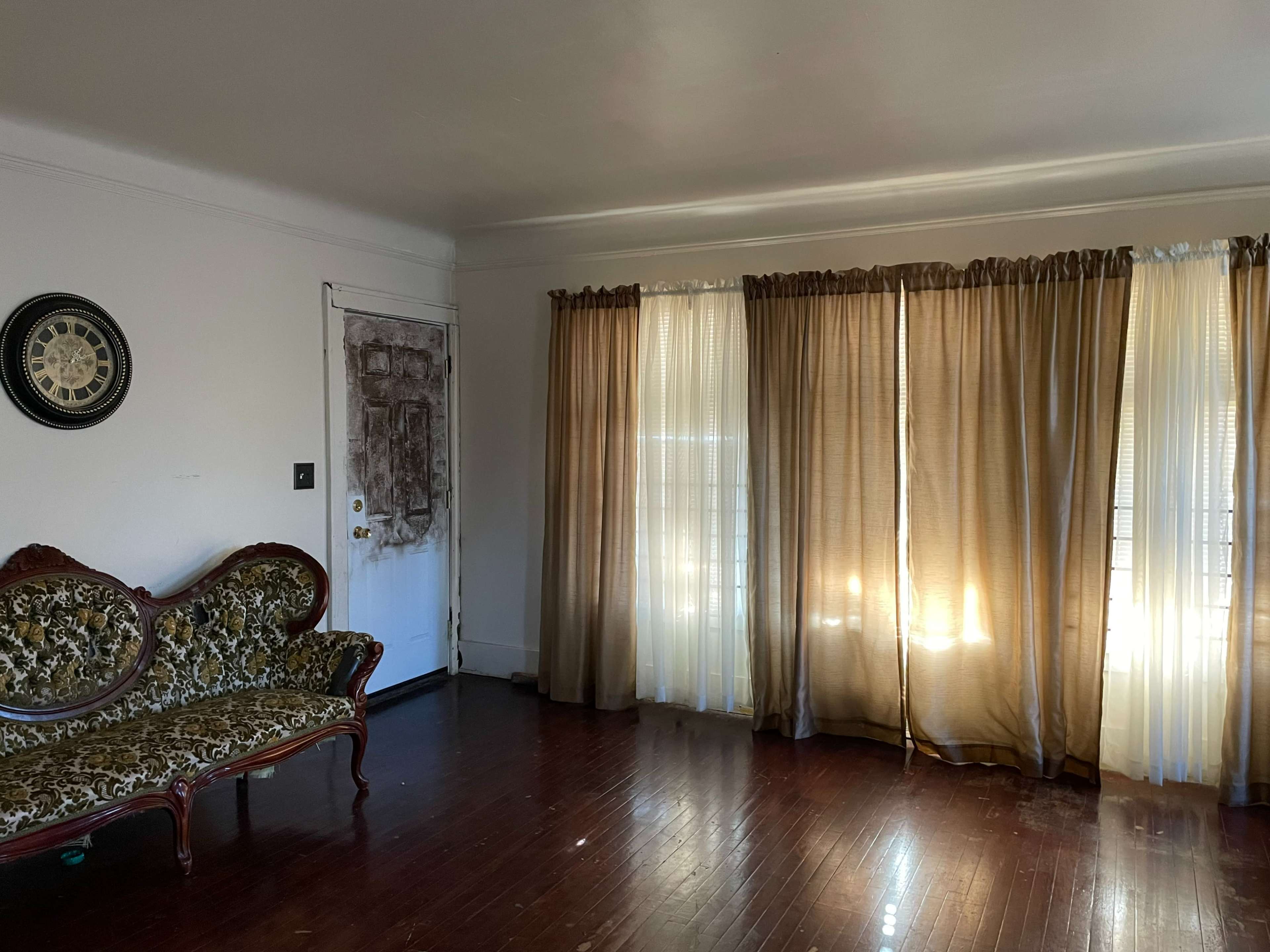 An empty room with a patterned couch, a weathered door, and light-colored curtains allowing sunlight to filter in.