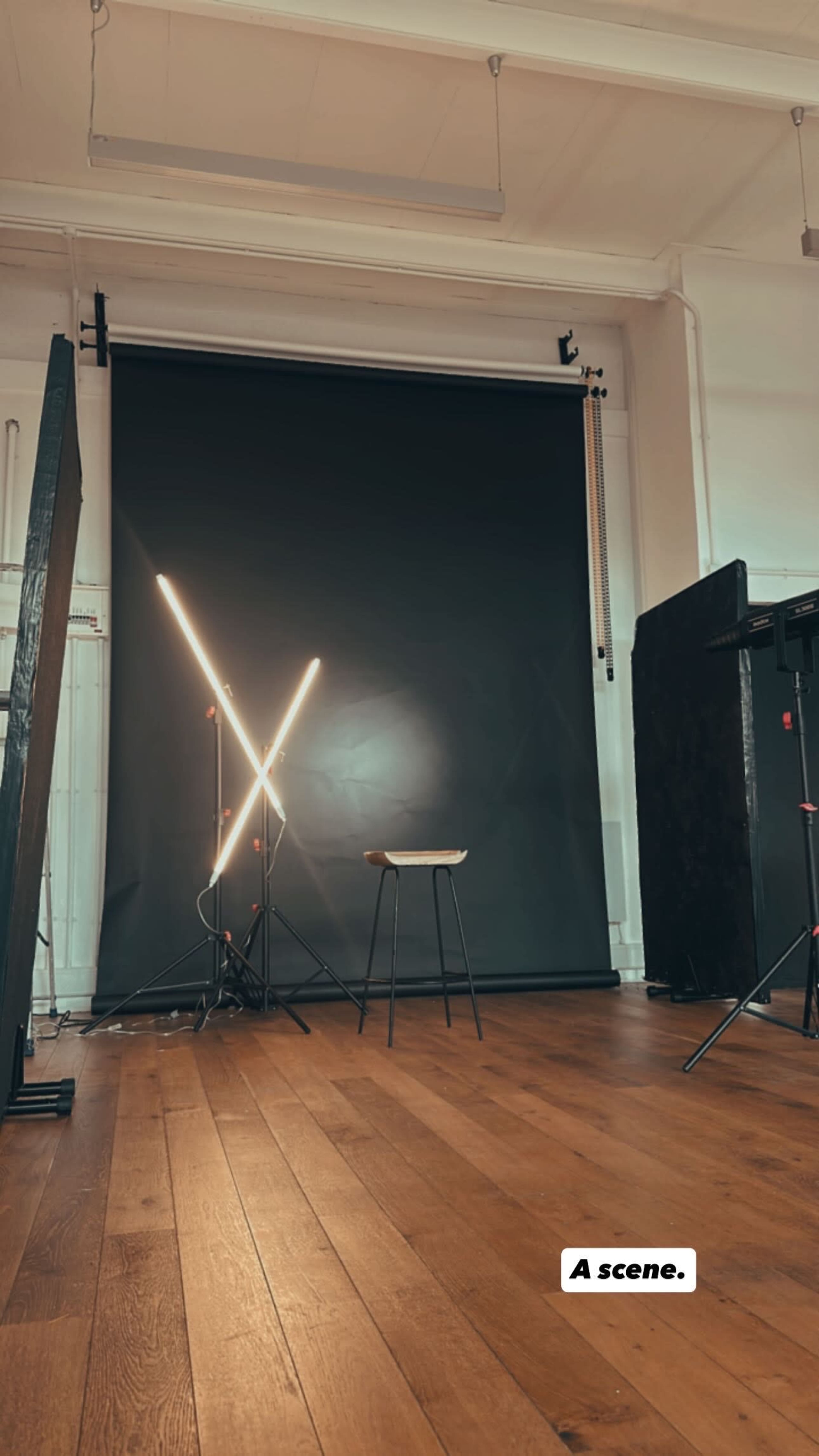 The image shows a photography studio with a dark backdrop, a wooden floor, and a stool positioned in front of LED lights arranged in an "X" shape.
