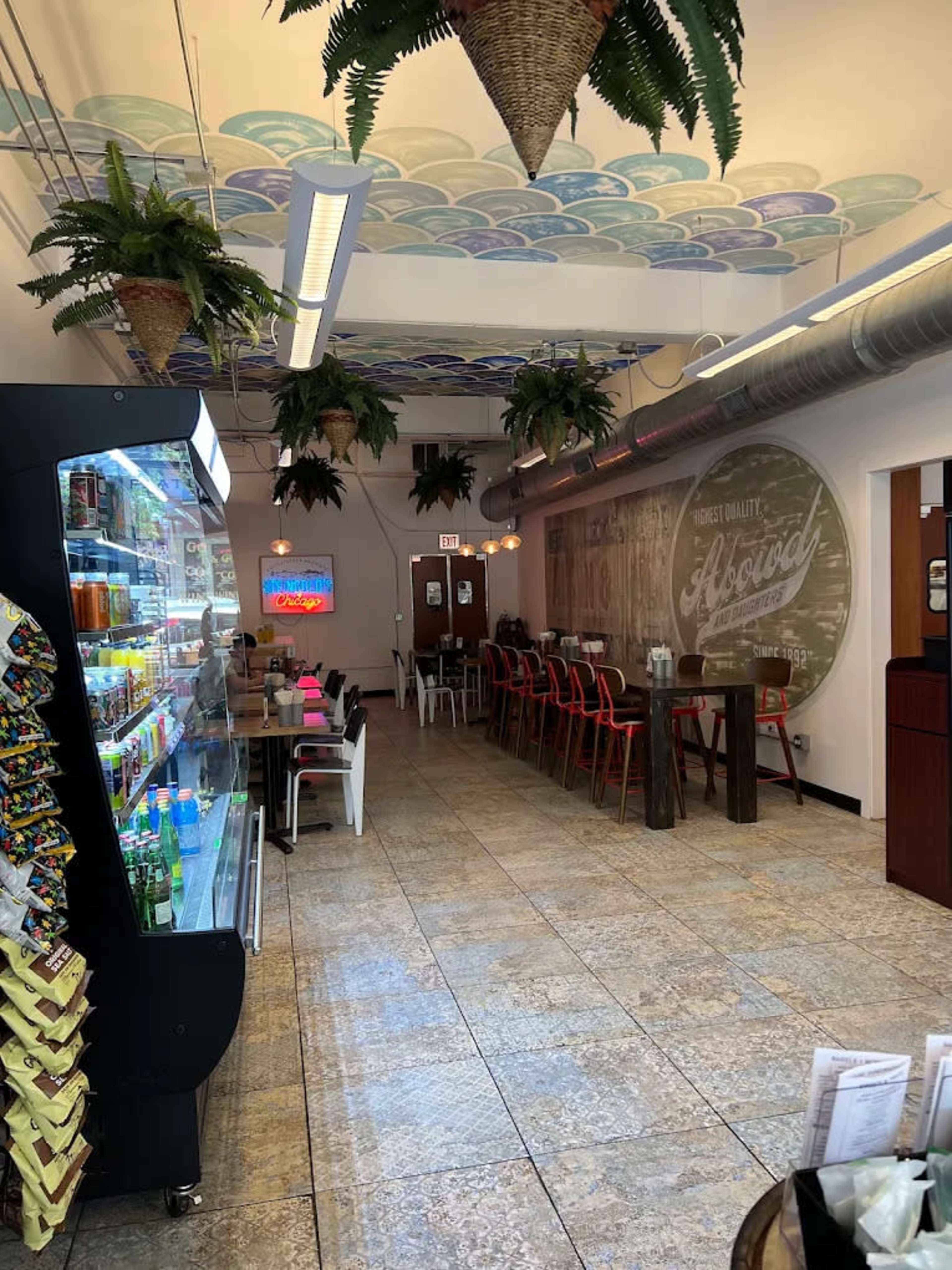 The image shows a cafe interior with a vending machine on the left, wooden tables and chairs arranged in the center, and decorative plants hanging from the ceiling.