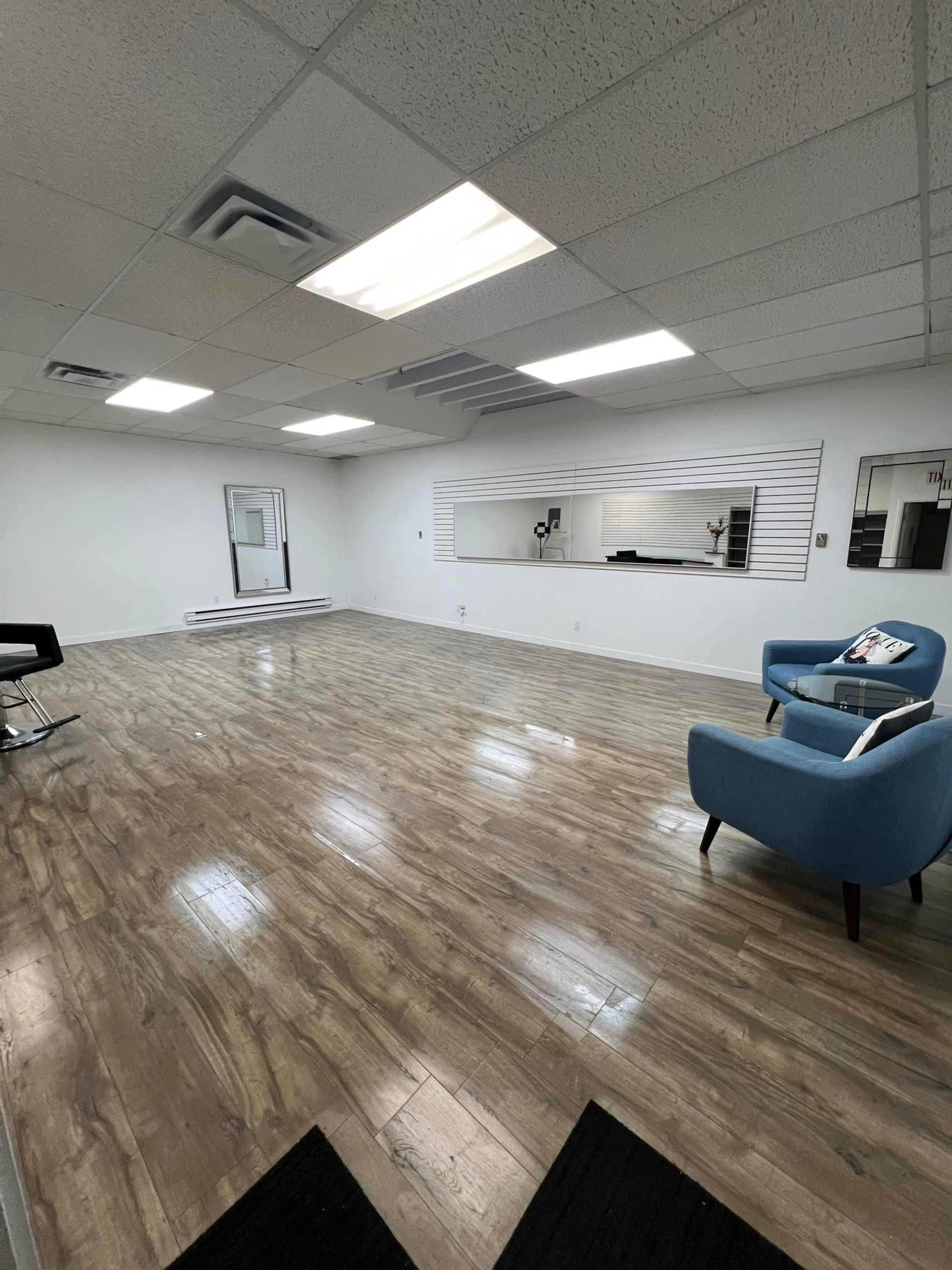 The image shows a spacious, empty room with wood-like flooring, featuring a blue armchair and a large mirror along one wall.